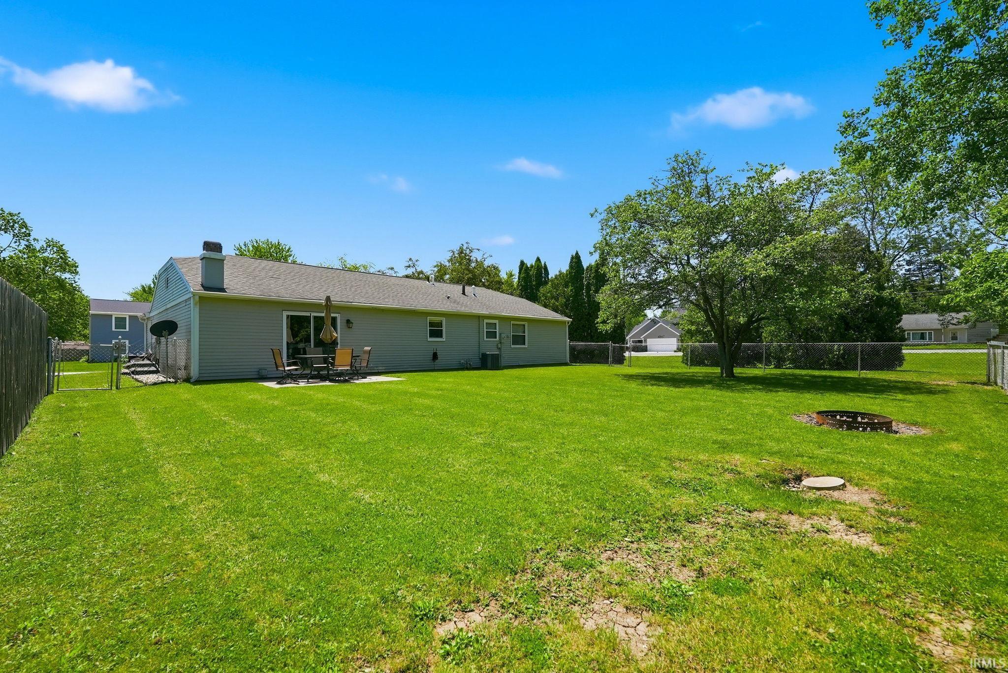 Expansive grassy backyard featuring a chain-link fence, mature trees, a patio with a sliding glass door, and a built-in fire pit