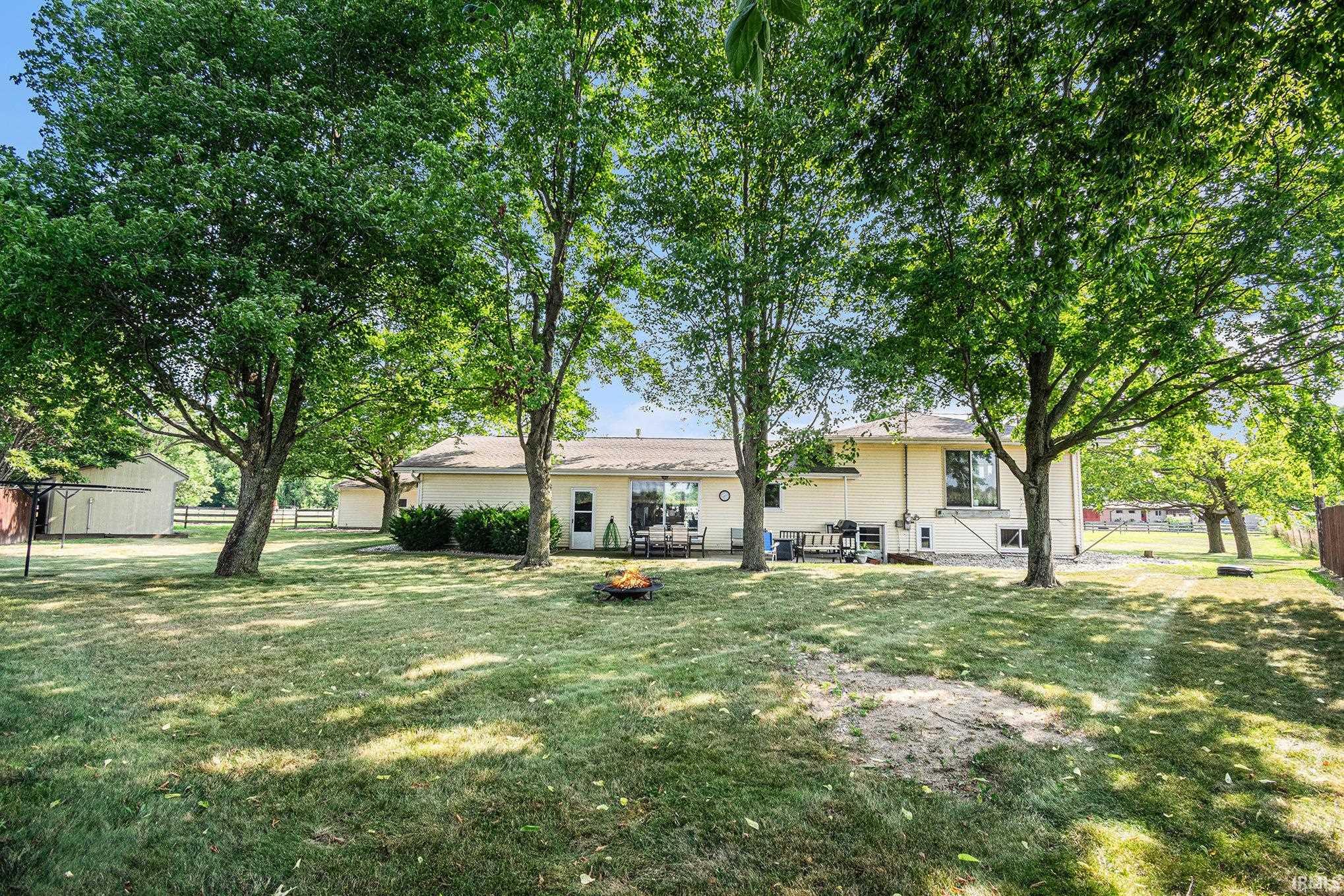 Expansive backyard featuring mature trees providing ample shade, a fire pit area, and a paved patio accessible via sliding glass doors