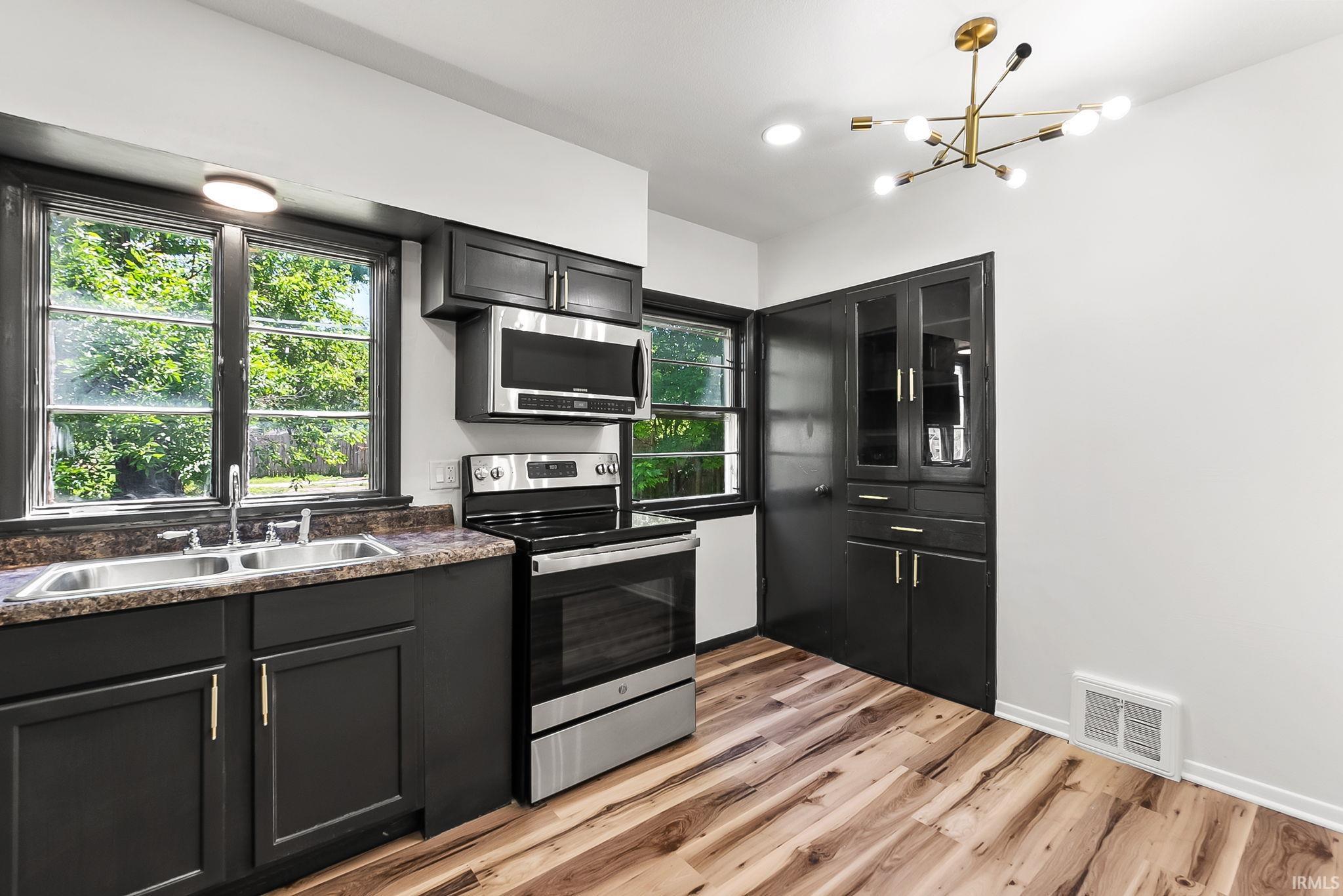 Kitchen featuring black cabinetry, stainless steel appliances, wood-finish flooring, and a modern light fixture