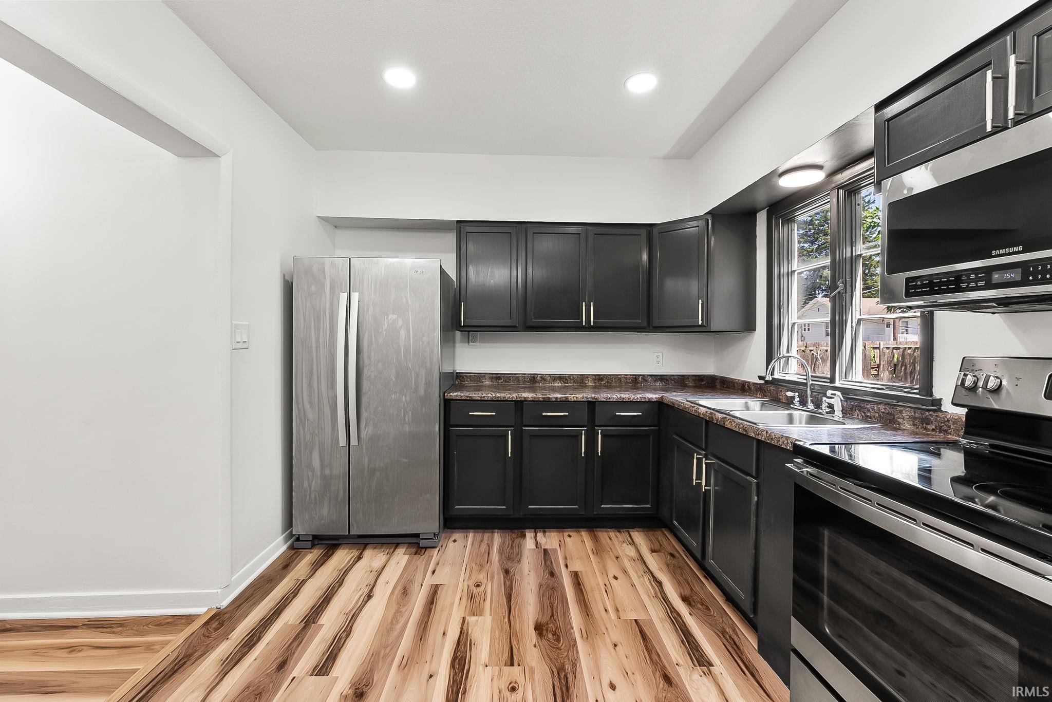 Kitchen featuring dark cabinetry, stainless steel appliances, wood-finish flooring, recessed lighting, and a double basin sink