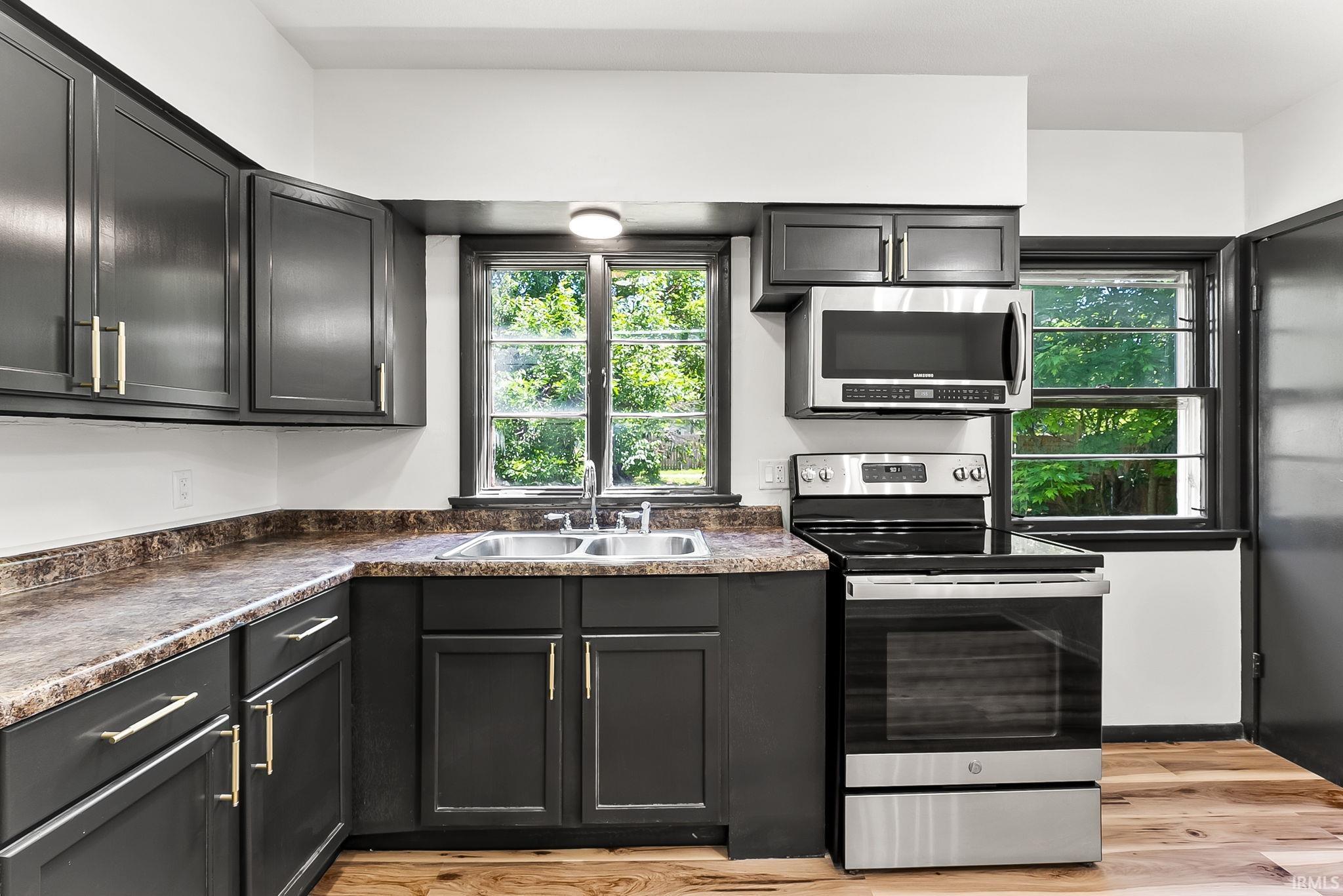 Kitchen featuring dark cabinetry with brushed gold hardware, a stainless steel electric range, an over-the-range microwave, a double basin sink, and wood-finish flooring