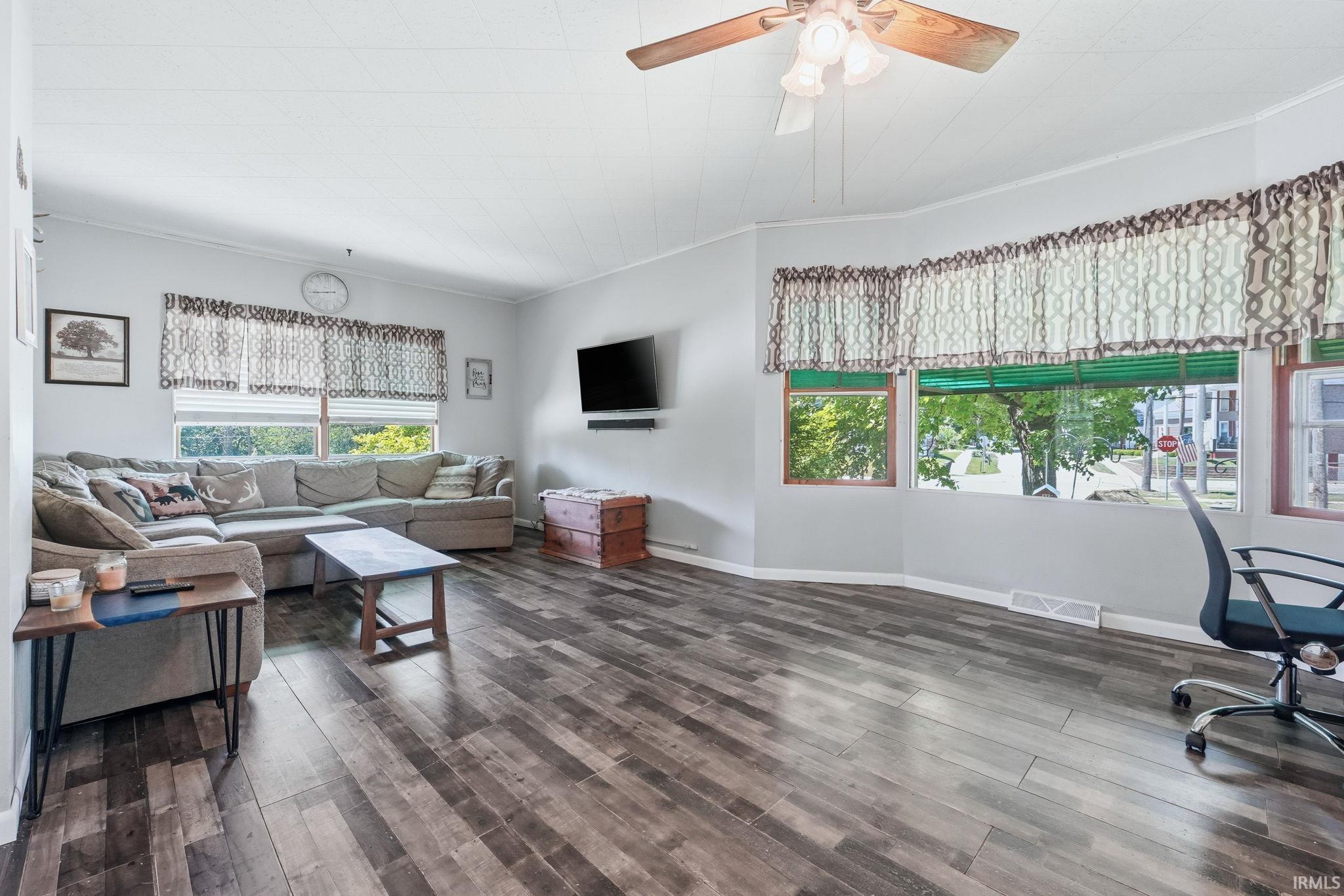 Spacious living area featuring wood-finish flooring, a ceiling fan with light fixture, and multiple large windows for natural light