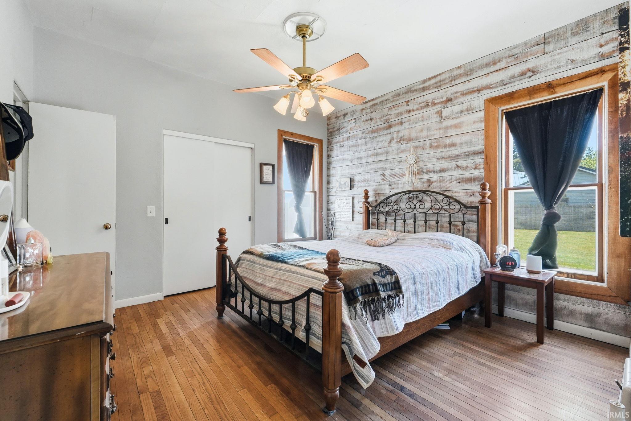 Spacious bedroom featuring wood-finish flooring, a prominent accent wall with horizontal wood paneling, and two windows with natural wood trim