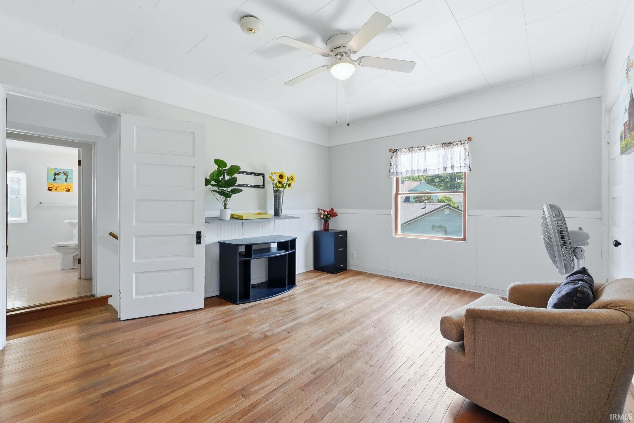 Hardwood flooring throughout, featuring a five-panel interior door, beadboard wainscoting, and a ceiling fan with integrated lighting