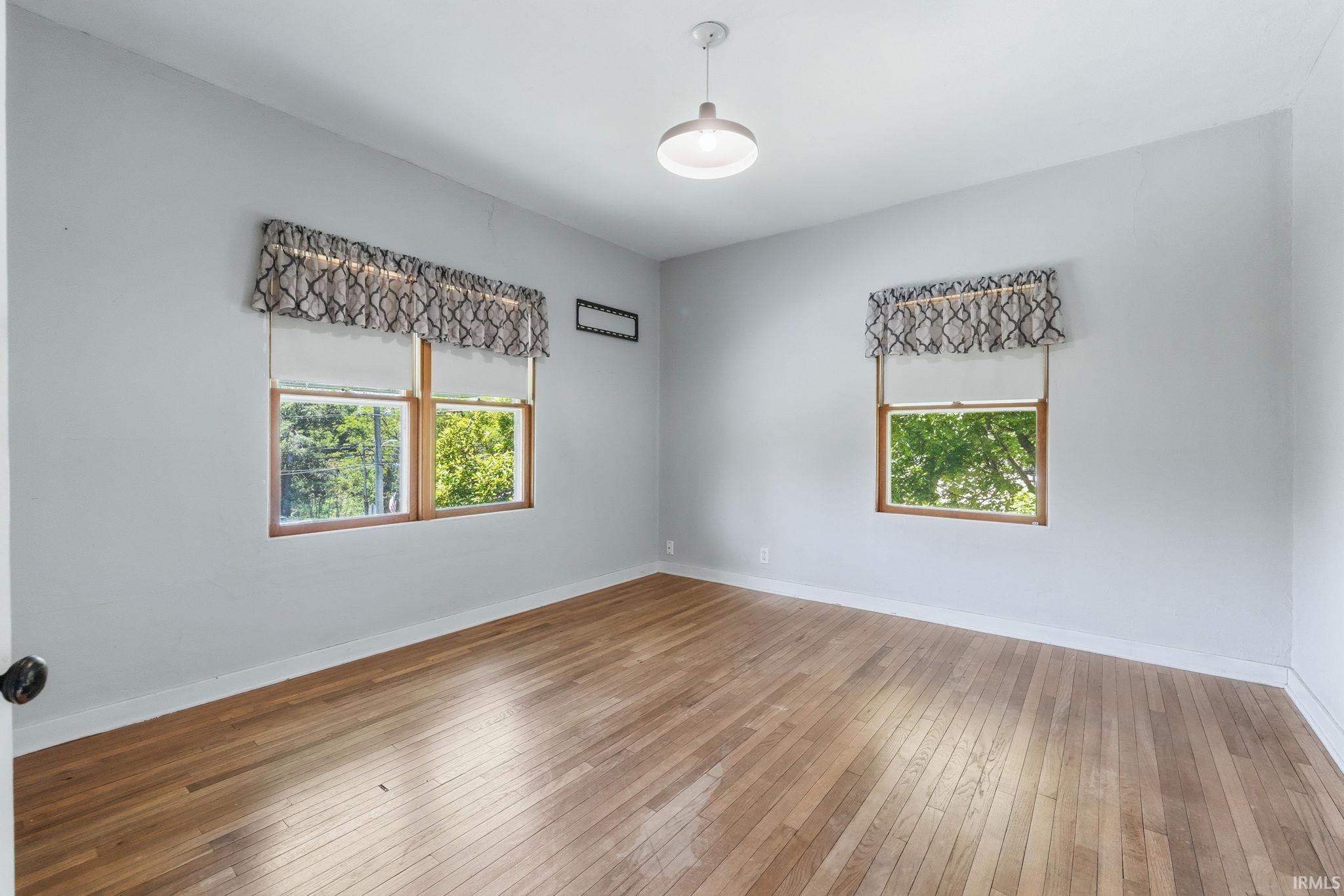 Light-filled room featuring wood-finish flooring, two windows with wooden frames, white baseboards, and a ceiling-mounted light fixture