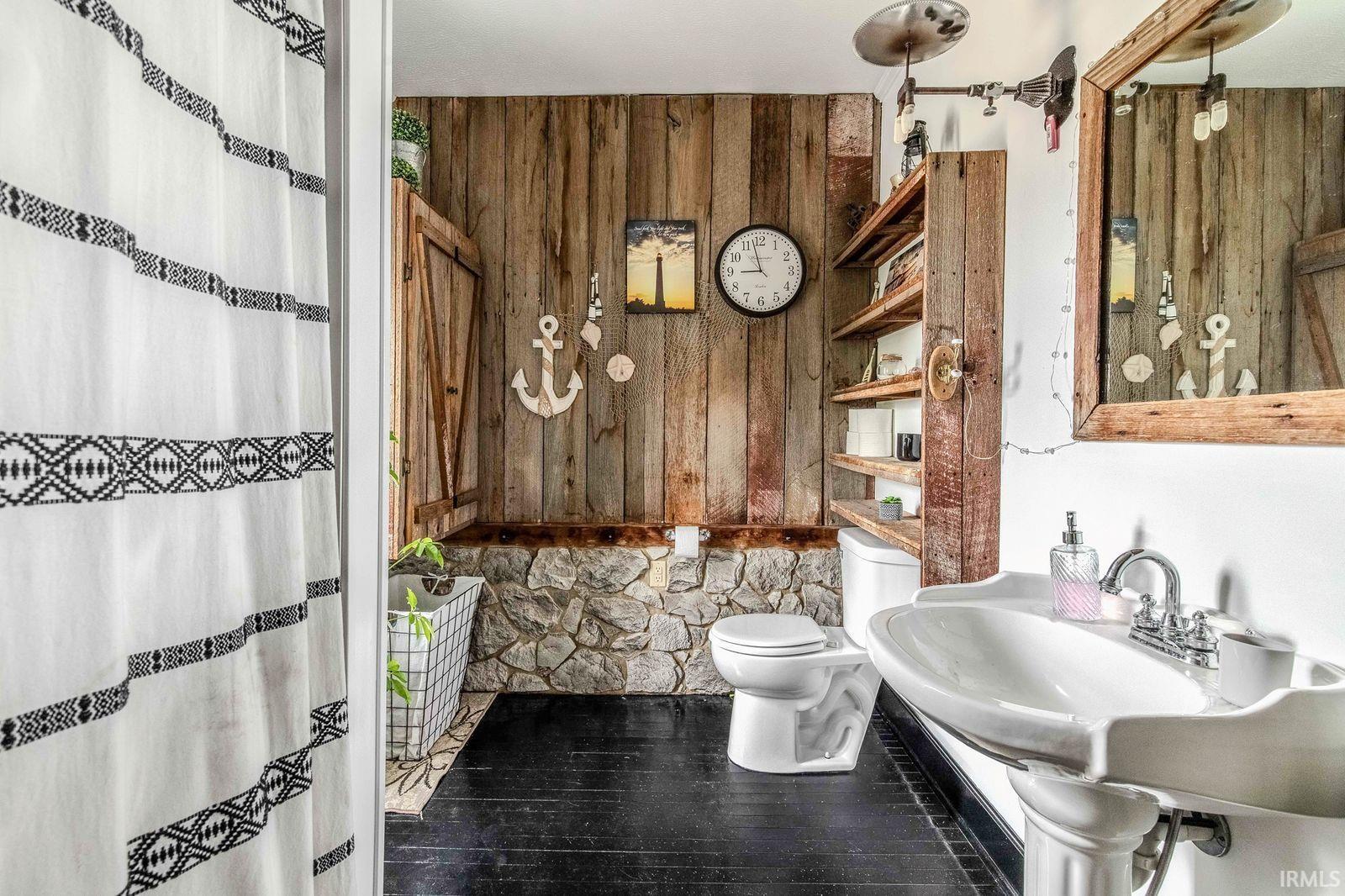 Bathroom featuring a pedestal sink with chrome fixtures, black flooring, stone and wood-paneled walls, a built-in wood shelving unit, and a white toilet