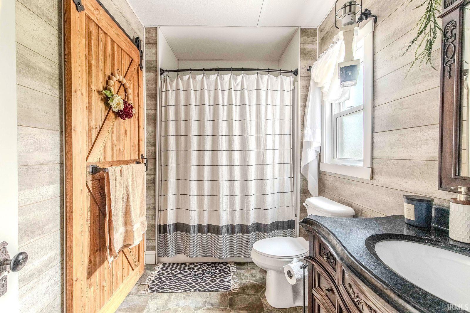 Bathroom featuring a sliding barn door, plank-style wall finishes, a built-in shower with a shower curtain, a single-pane window, and a vanity with an integrated sink and dark countertop