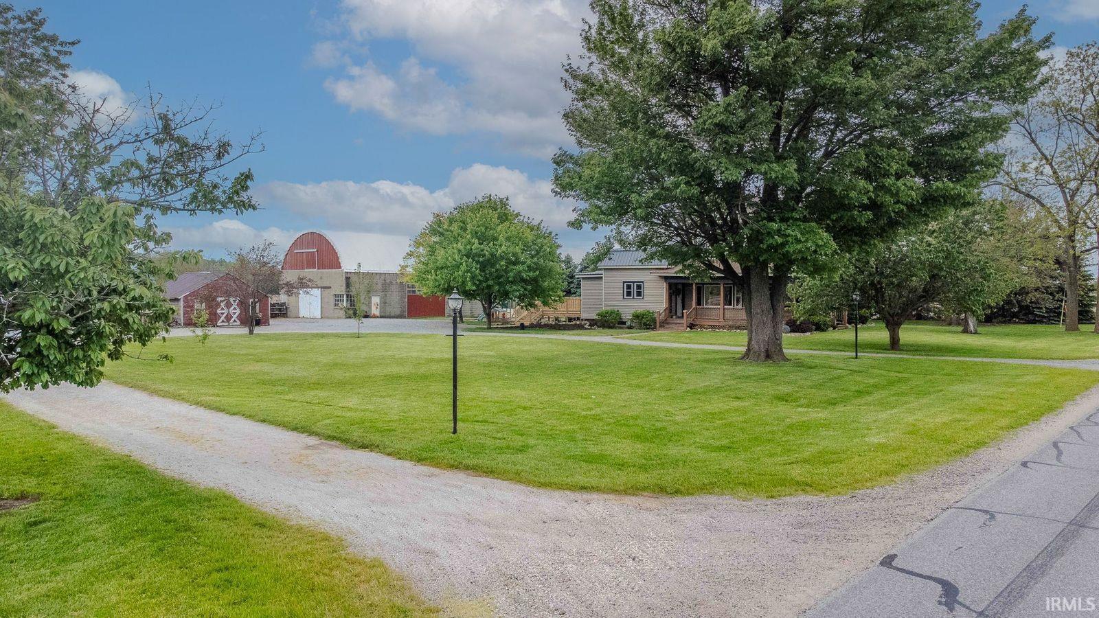 Expansive property featuring a gravel driveway, mature trees, manicured lawns, a single-story residence with a covered porch, and multiple auxiliary structures including a red barn