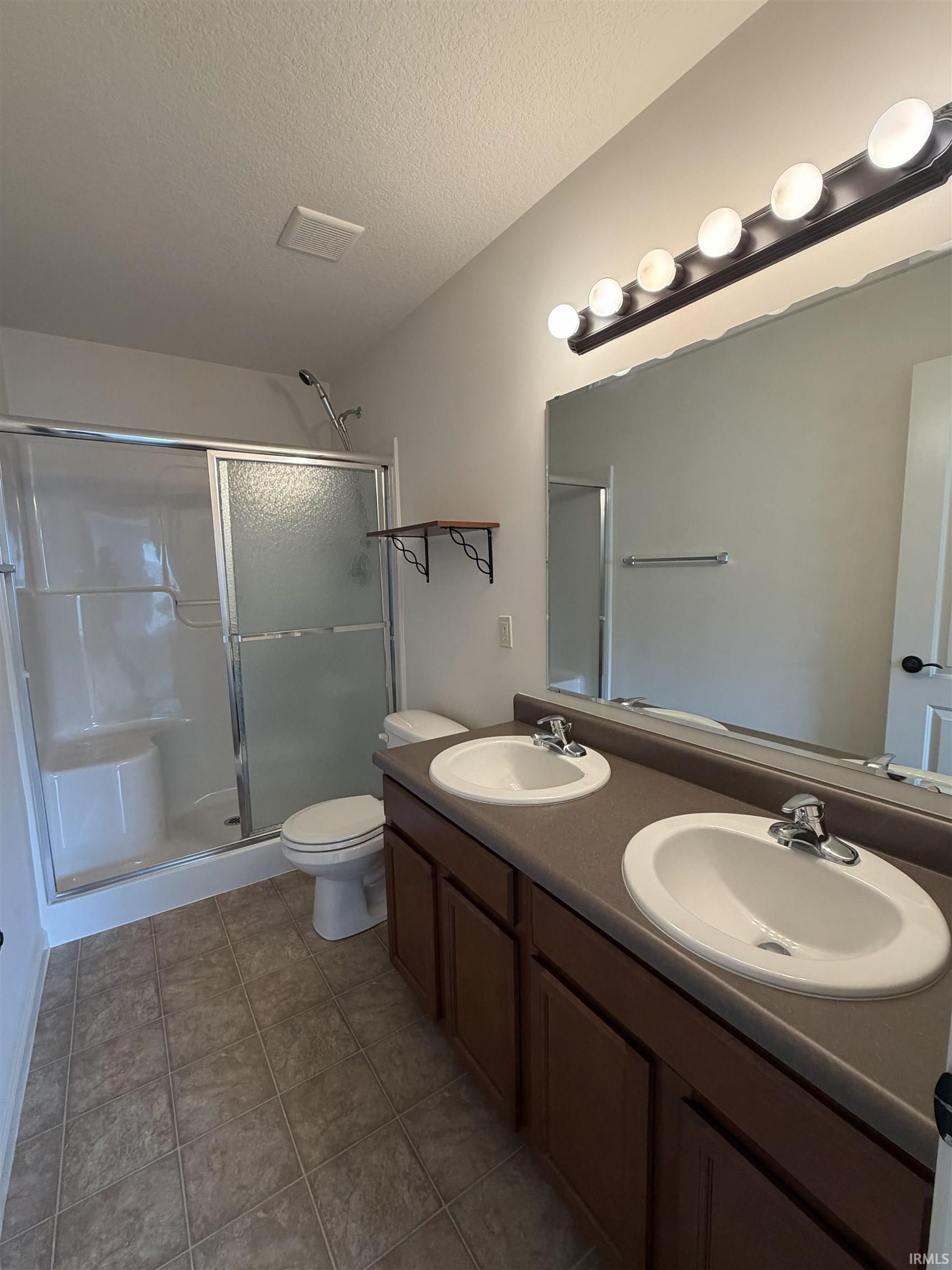 Bathroom featuring a dual vanity with a dark countertop and white oval sinks, a large wall-mounted mirror, a shower with a sliding glass door and integrated seating, and tiled flooring