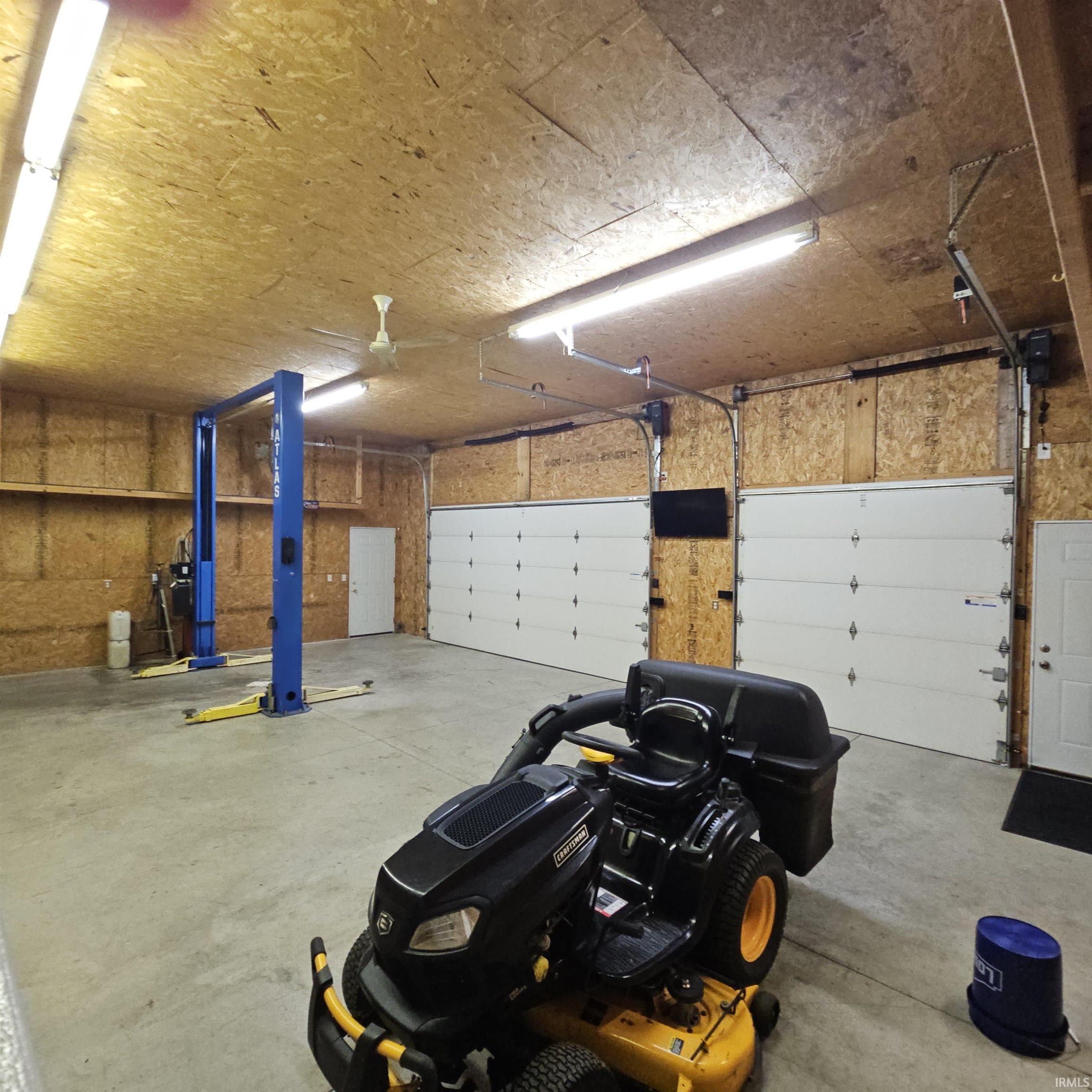 Spacious garage featuring a concrete floor, two white overhead doors, exposed wood-finish walls, overhead fluorescent lighting, and a ceiling fan