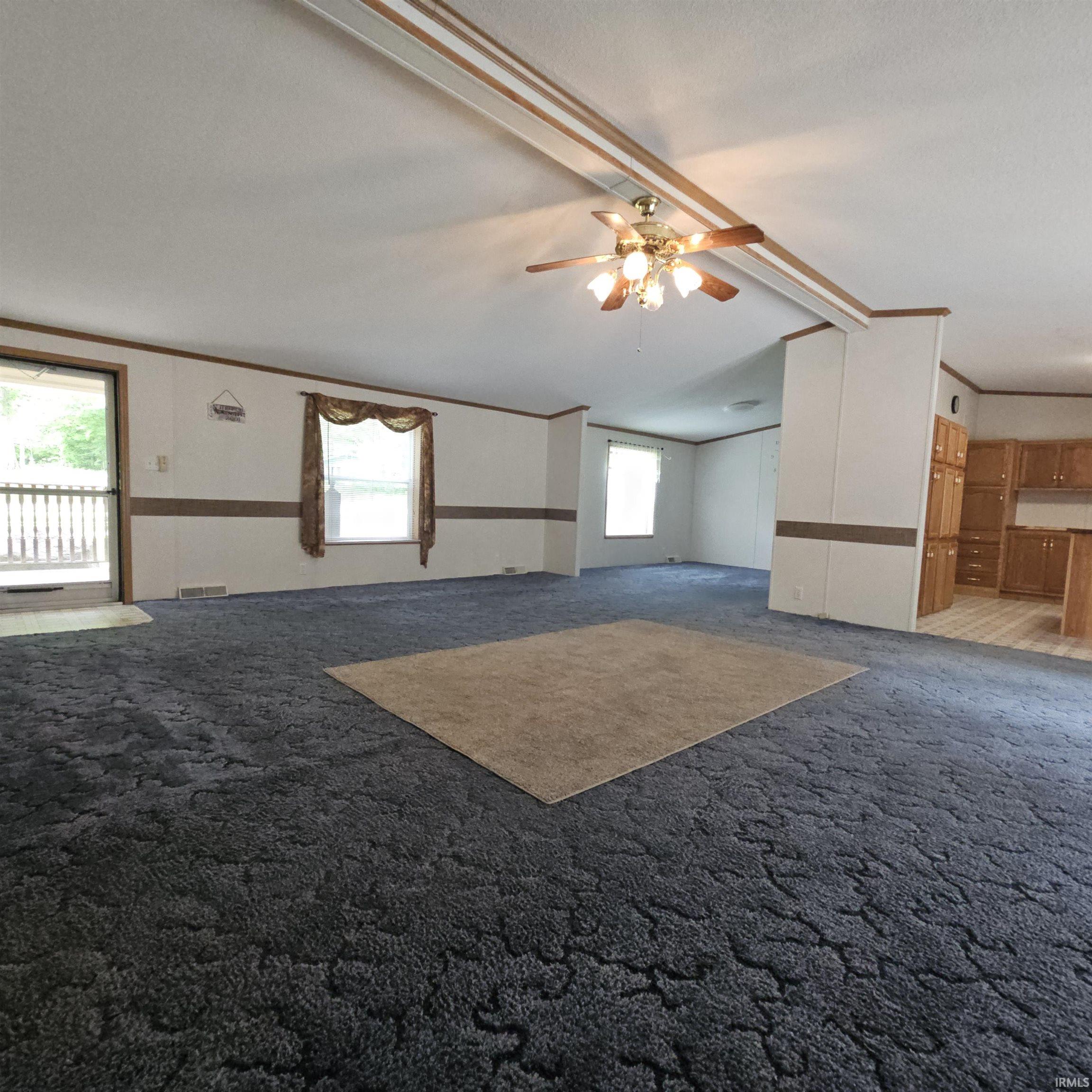 Spacious living area featuring patterned blue carpeting, white walls with a brown accent band, a ceiling fan with light fixture, and multiple windows providing natural light