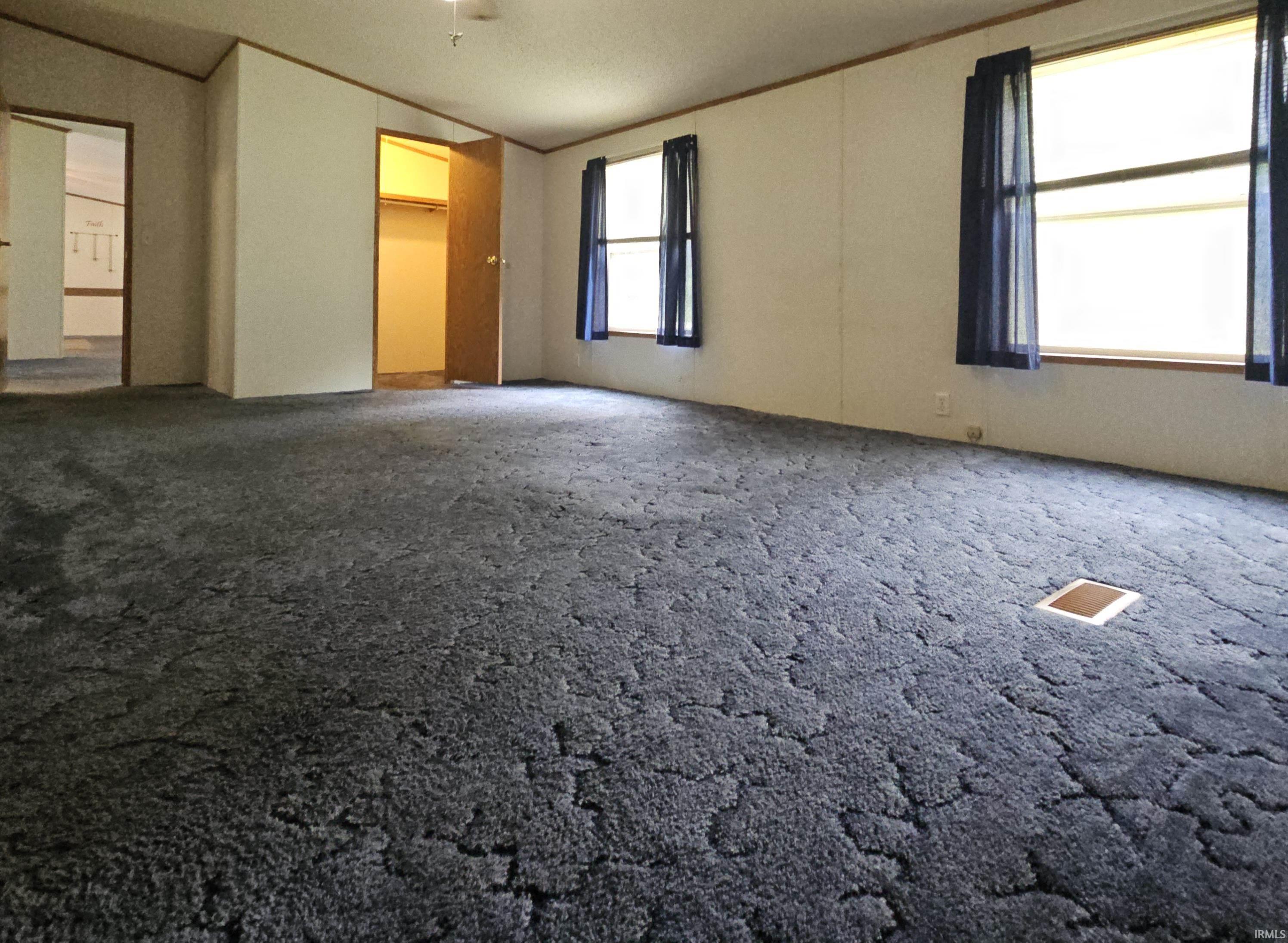 Spacious interior room featuring textured carpeting, multiple windows, white paneled walls, and a recessed floor outlet