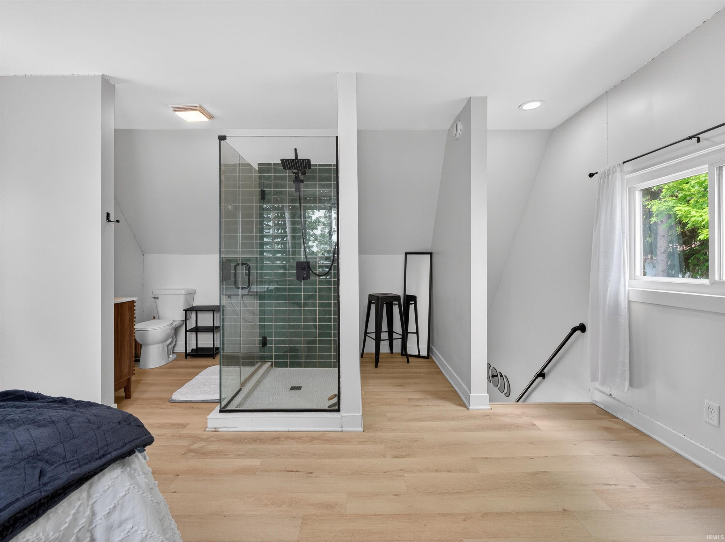 Open-concept bathroom featuring a glass-enclosed shower with green subway tile, light wood-finish flooring, a white toilet, and a white-framed window