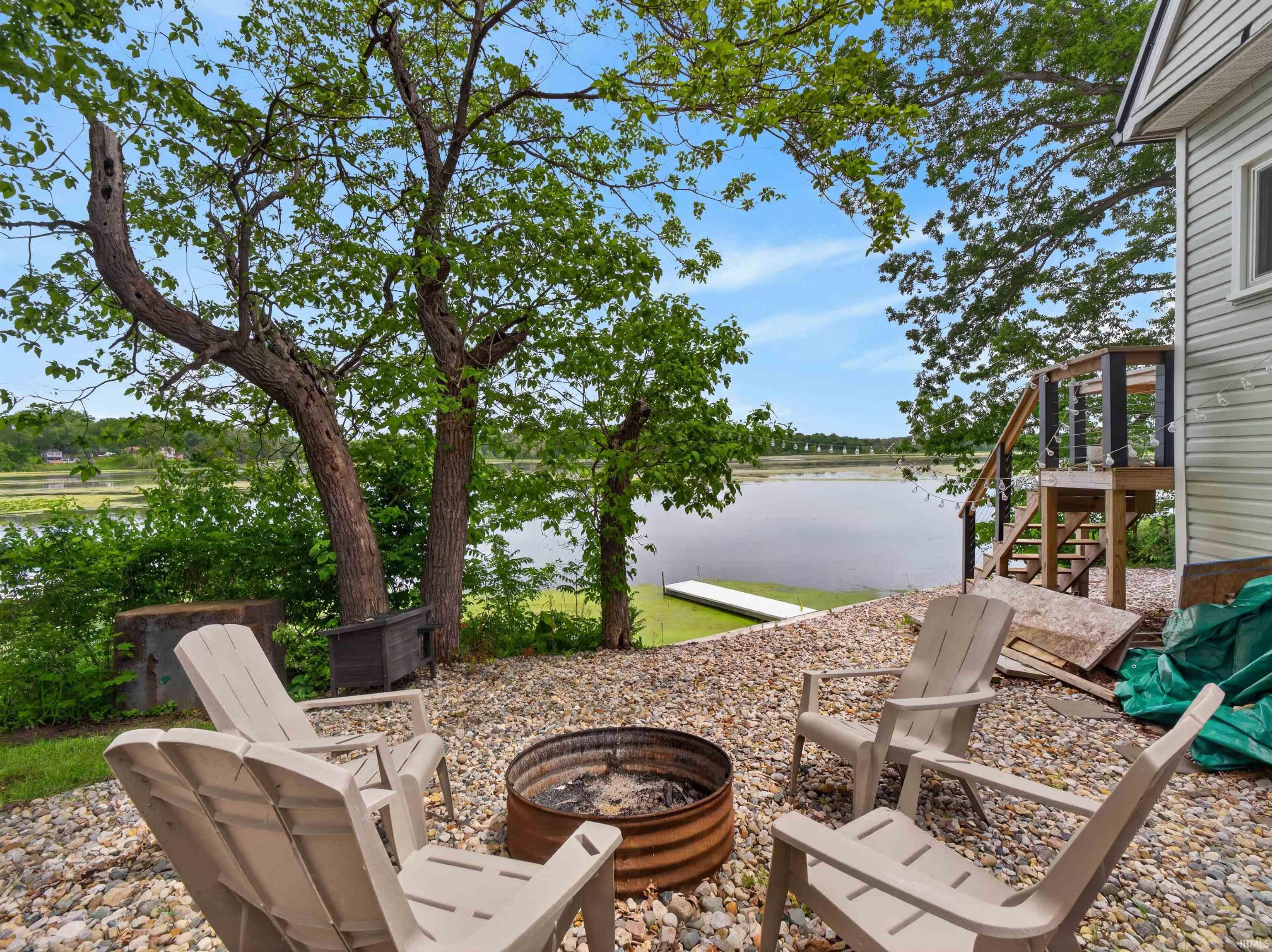 Gravel patio area featuring a built-in fire pit, mature trees, and waterfront access with a dock