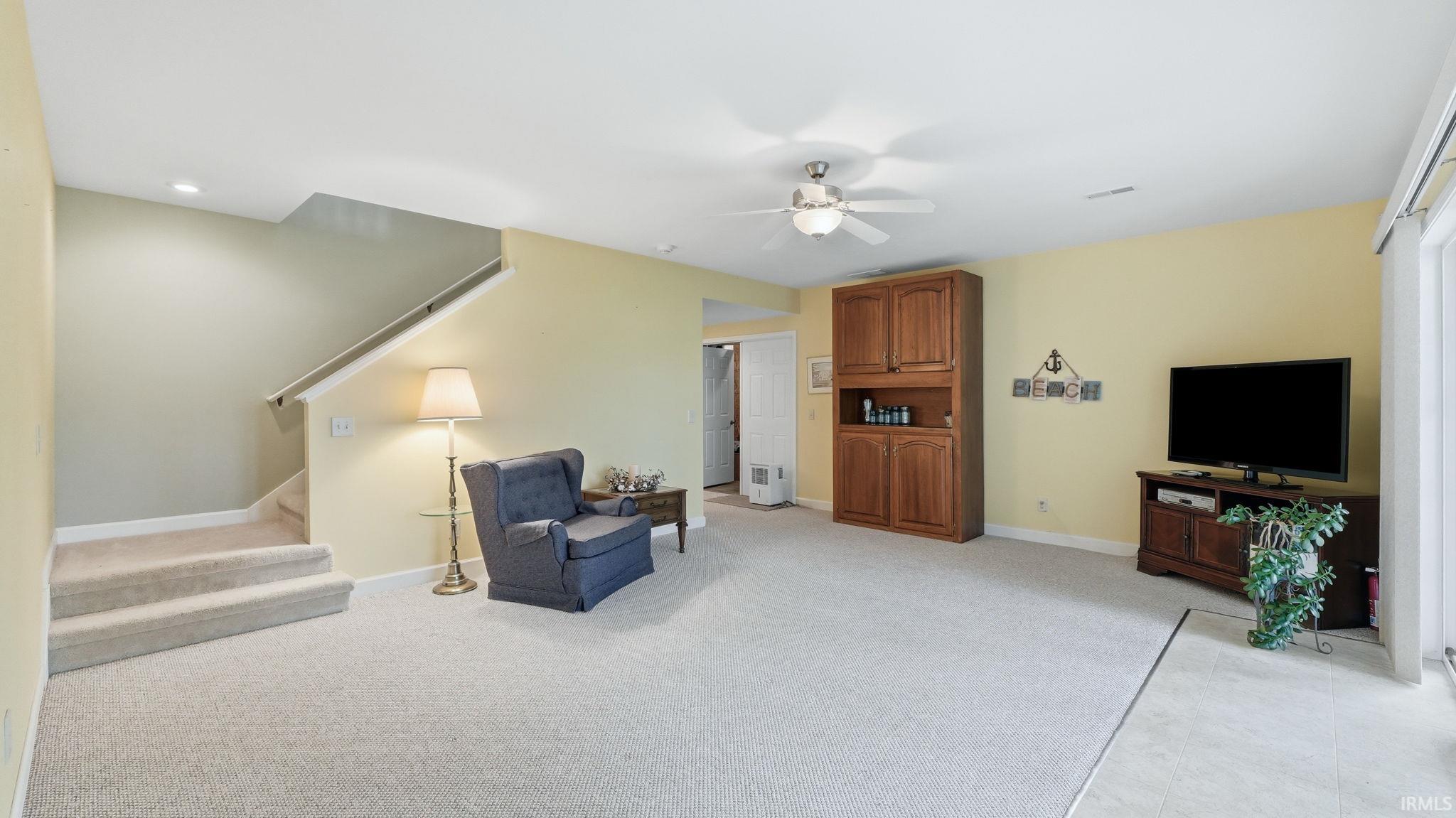 Carpeted lower-level living area featuring a wood-finish built-in cabinet, ceiling fan with integrated lighting, and a white-trimmed staircase with carpeted steps