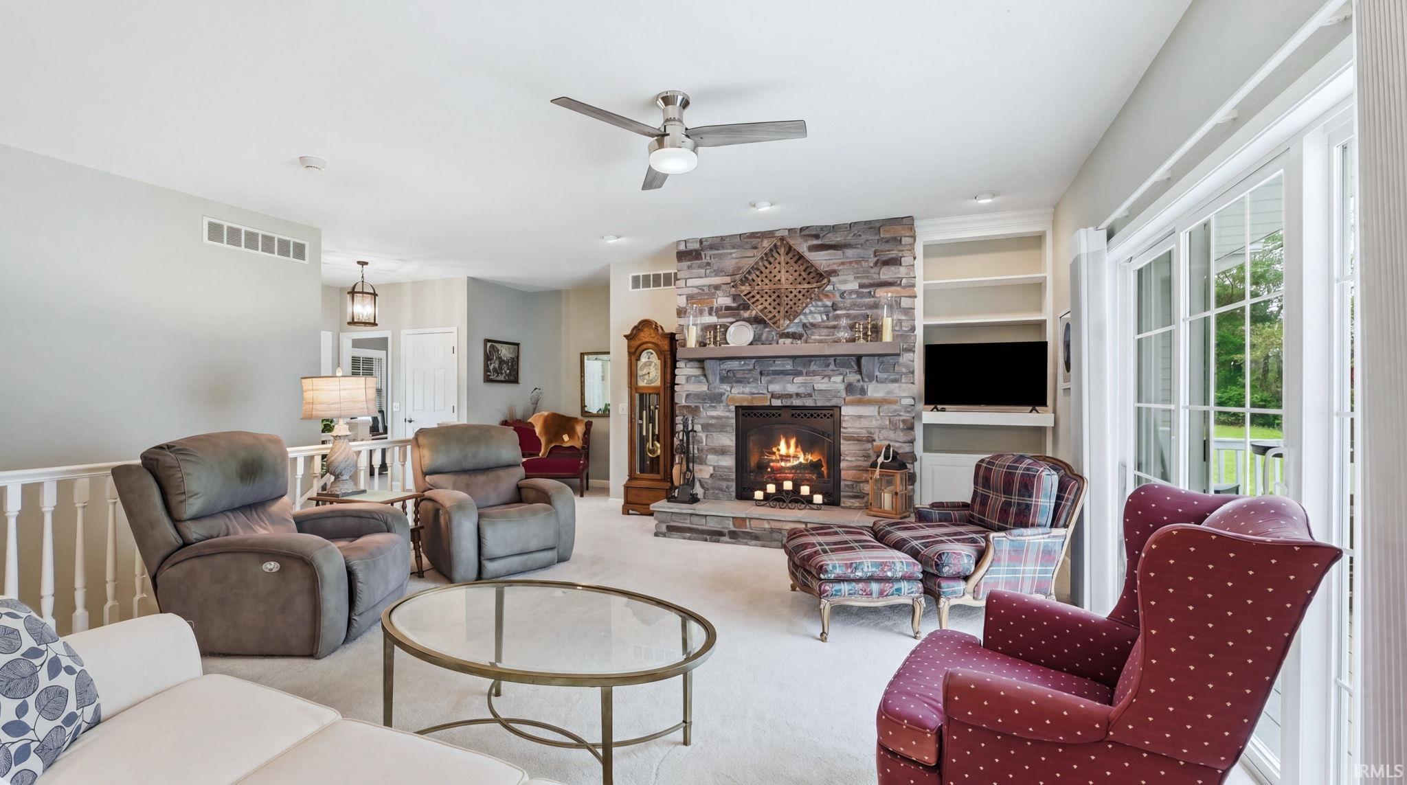 Living space featuring a stone-clad fireplace with a wood mantel, built-in shelving, glass sliding doors, a ceiling fan with integrated lighting, and neutral carpet flooring
