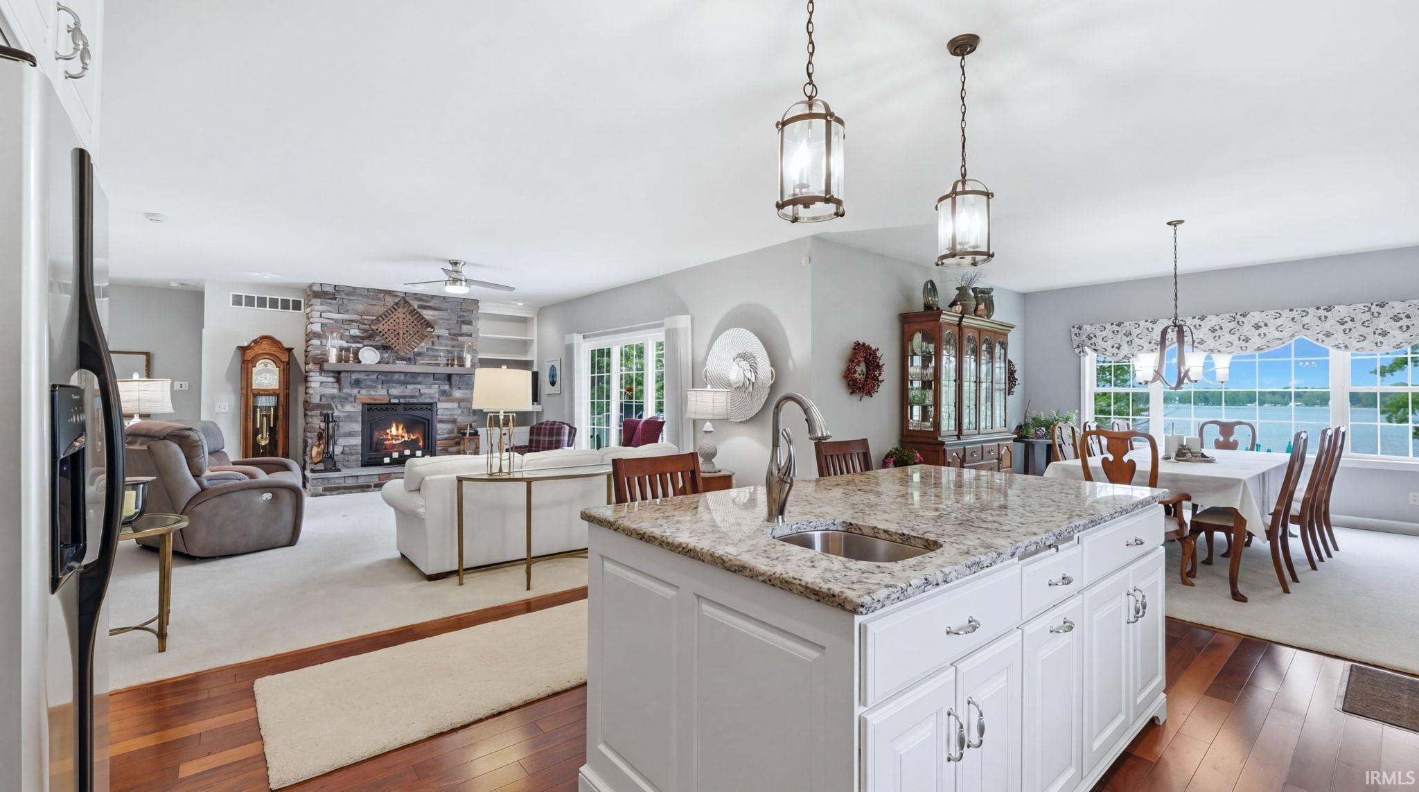 Open concept living area with a stone-clad fireplace, built-in shelving, wood-finish flooring, a kitchen island with a granite countertop and integrated sink, and an expansive window overlooking a body of water