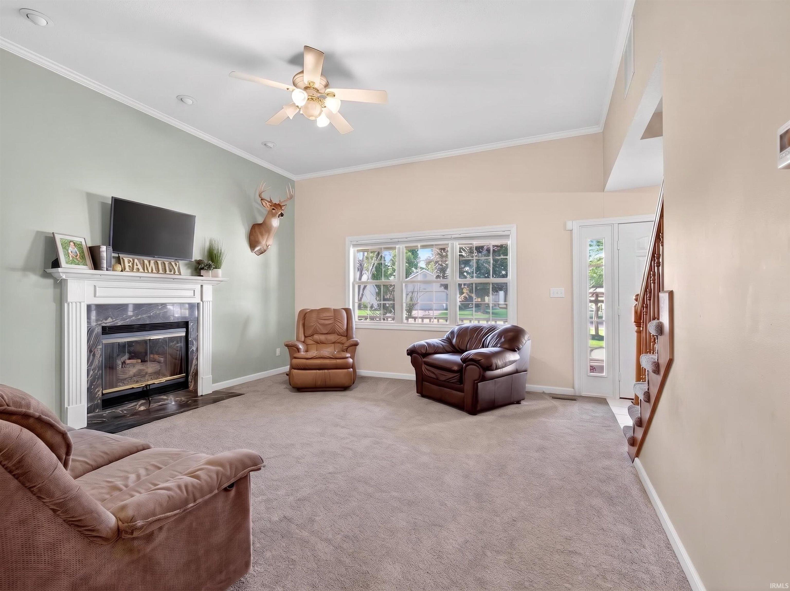 Living room with vaulted ceiling featuring a white mantel fireplace with a dark stone surround, a ceiling fan with integrated lighting, and neutral carpeting