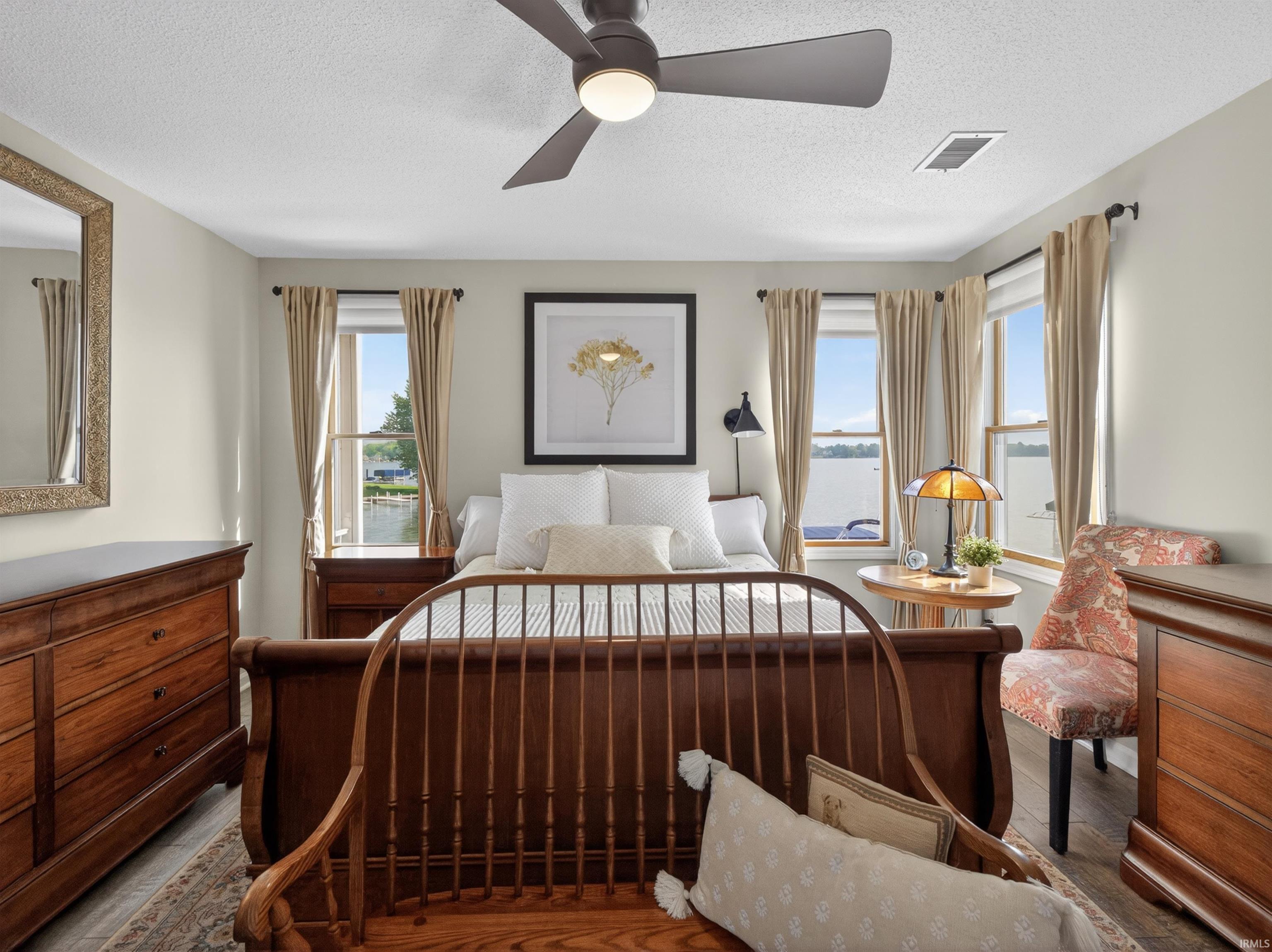 Spacious bedroom featuring multiple windows offering water views, wood-finish flooring, a ceiling fan with integrated lighting, and neutral wall tones