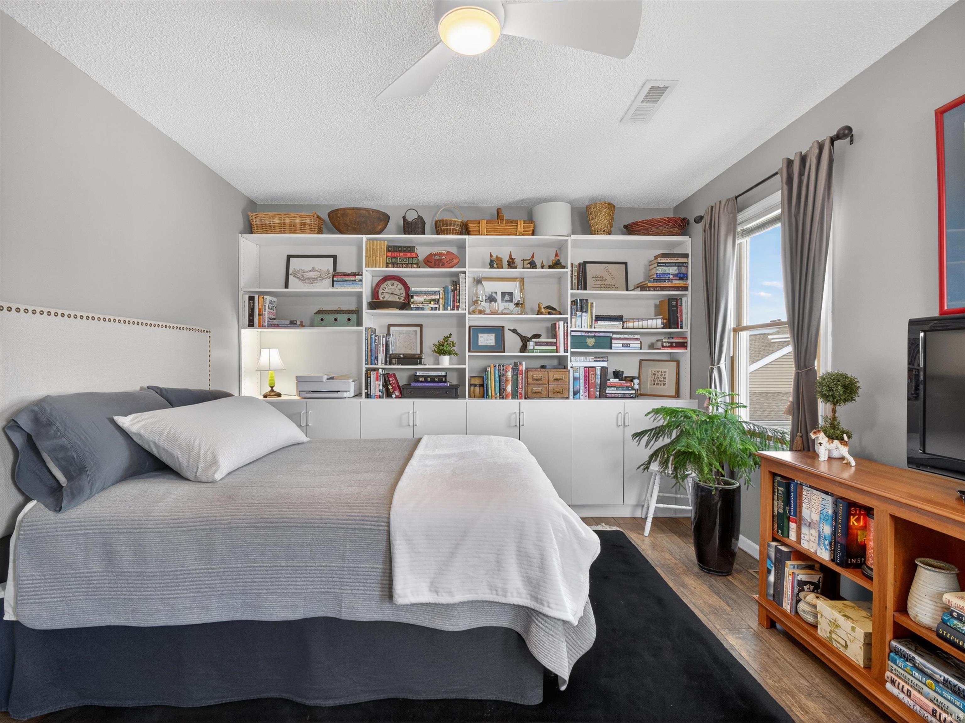 Spacious room featuring a wall-spanning built-in shelving unit, wood-finish flooring, a ceiling fan, and a large window with grey drapes