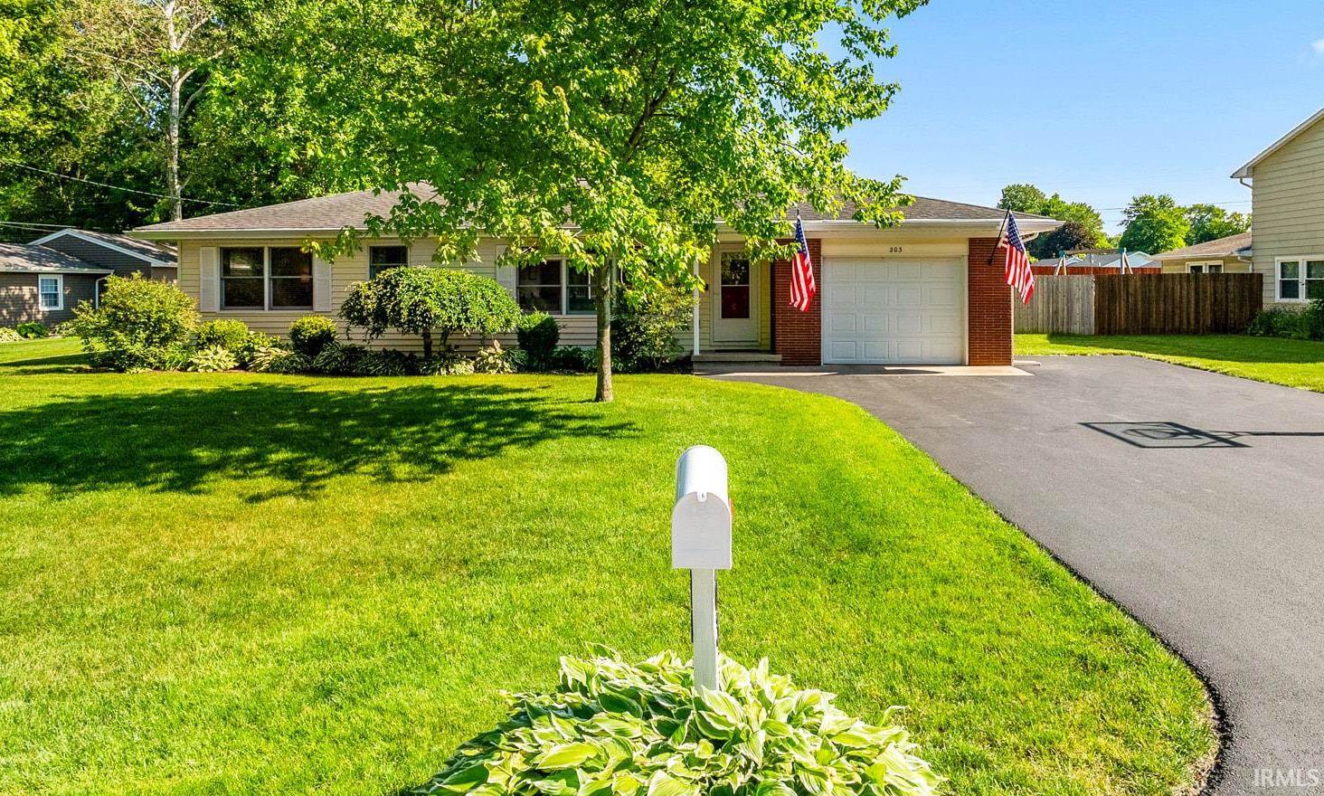 Single-story residence featuring a front-facing garage with a white door, a paved driveway, and mature trees providing natural shade