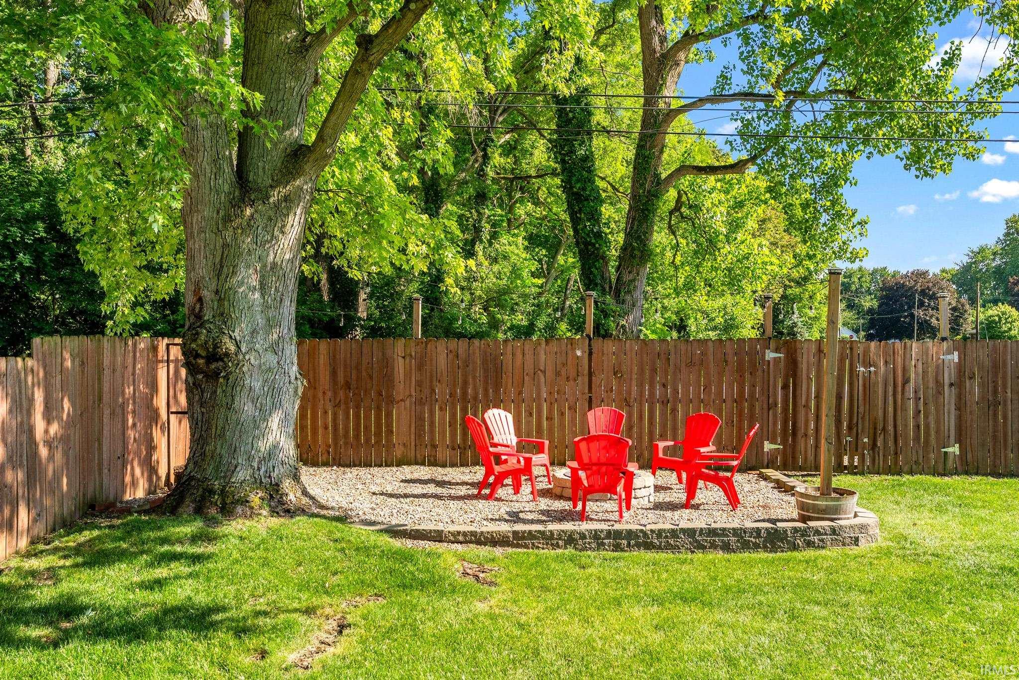 Expansive backyard featuring a stone-lined fire pit area with gravel ground cover, a natural wood privacy fence, and mature trees providing shade