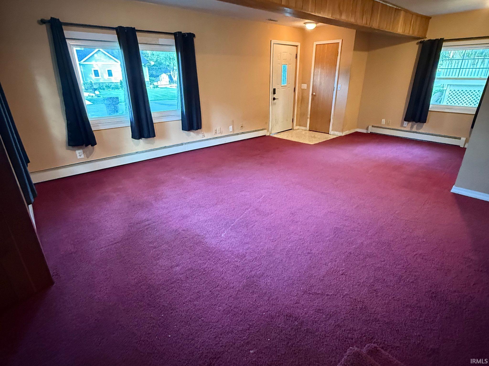 Spacious room featuring rich purple carpeting, multiple windows, a wood-paneled ceiling accent, and a front entry door with a frosted glass insert