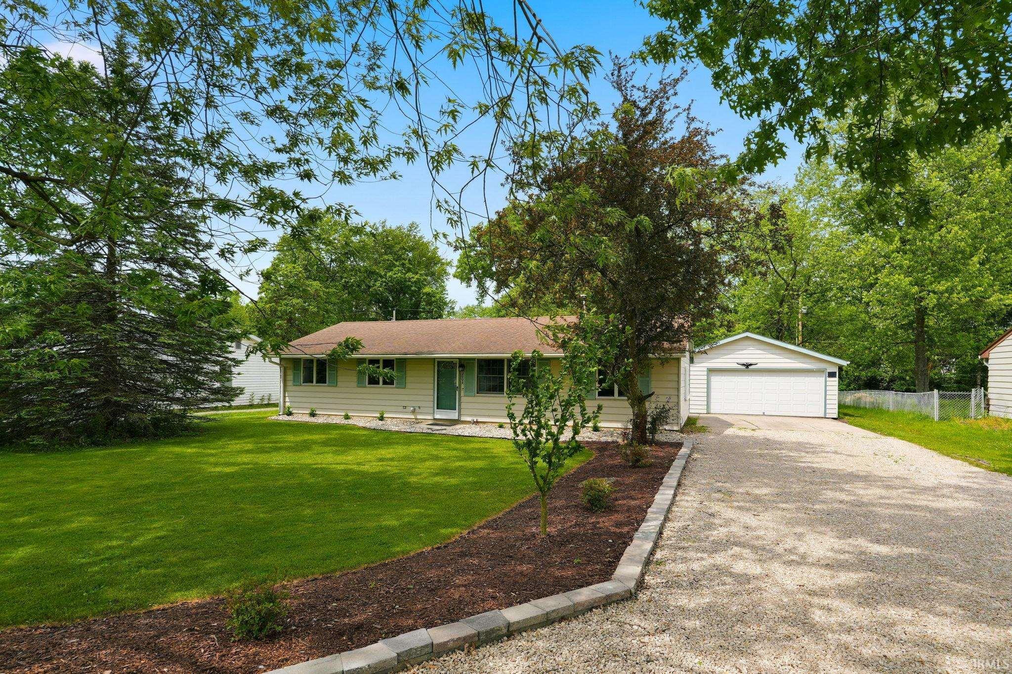 Ranch-style residence featuring a gravel driveway, an attached two-car garage, mature trees, and a landscaped front yard with a mulch bed