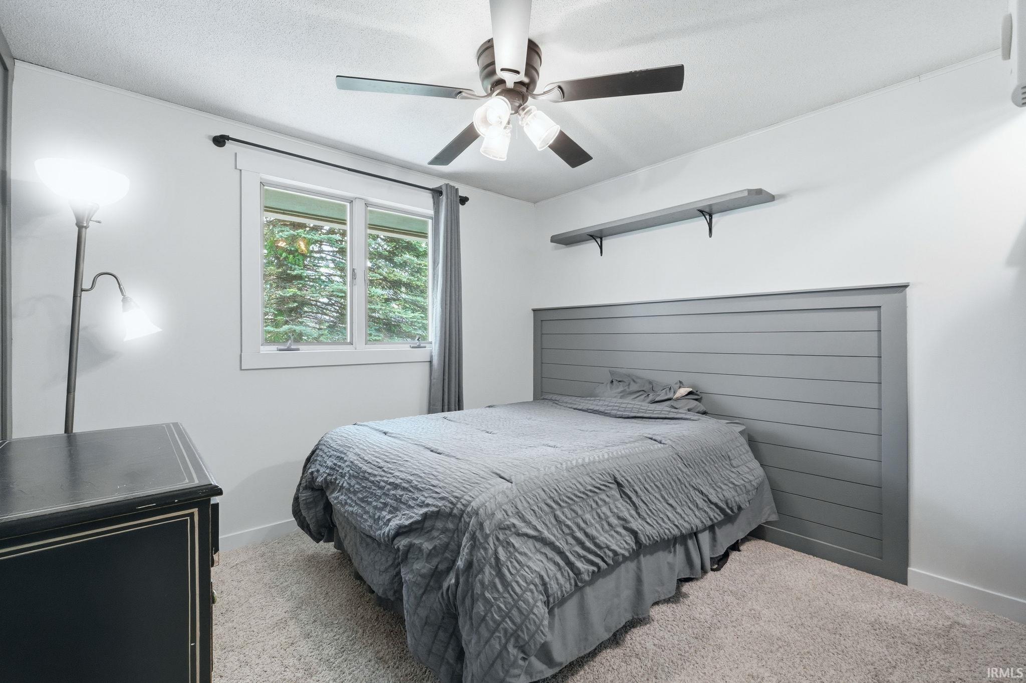 Bedroom featuring a ceiling fan with light fixture, double-pane window, wall-mounted shelving, built-in headboard, and carpeted flooring