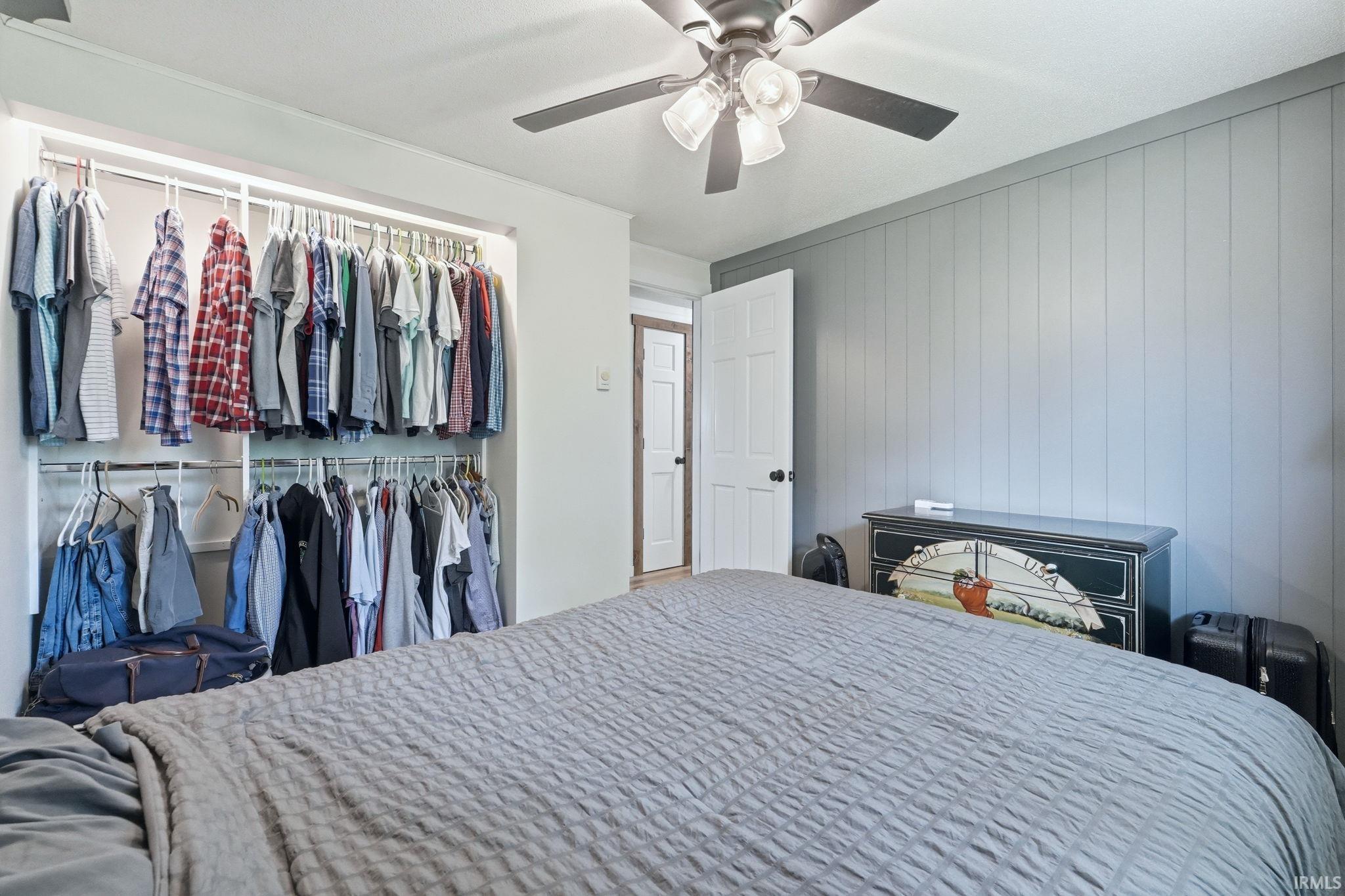 Bedroom featuring a ceiling fan with integrated lighting, a spacious open closet system with multiple hanging rods, and an accent wall with vertical paneling