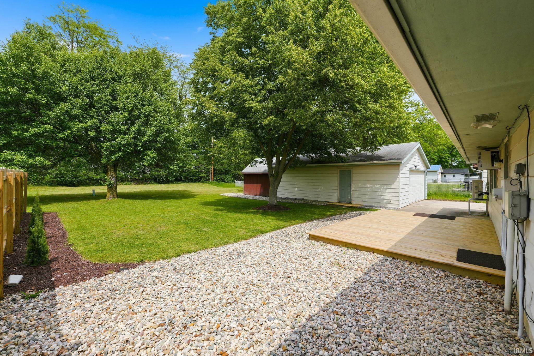 Rear exterior featuring a wooden deck, gravel patio, expansive lawn, mature trees, and a detached garage with siding