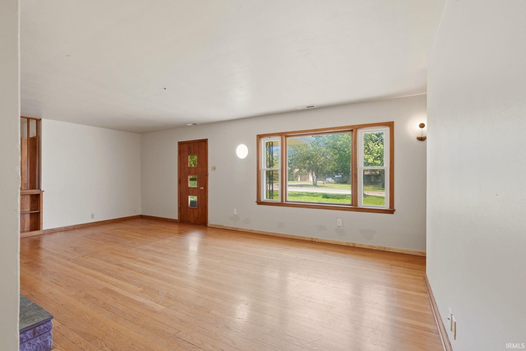 Hardwood flooring throughout a spacious room featuring a wood-panel entry door, large multi-pane windows with wood trim, and a built-in shelving unit