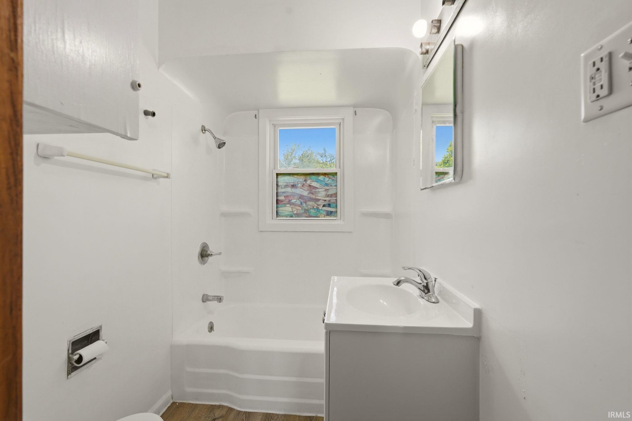 Bathroom featuring a white tub/shower combination, white vanity with integrated sink, chrome faucet, and wood-finish flooring