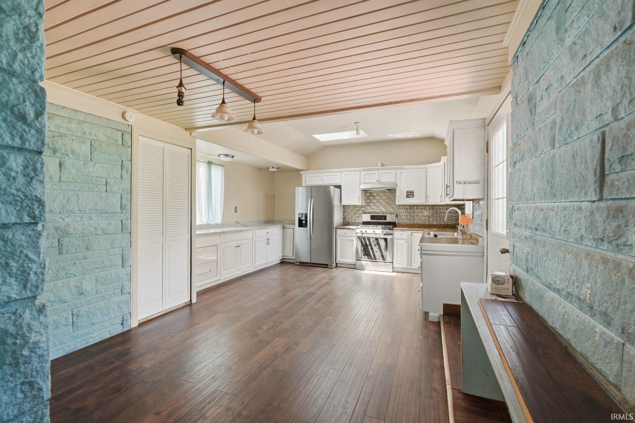 Spacious kitchen and dining area with wood-finish flooring, white cabinetry, integrated stainless steel appliances, a tile backsplash, and a skylight