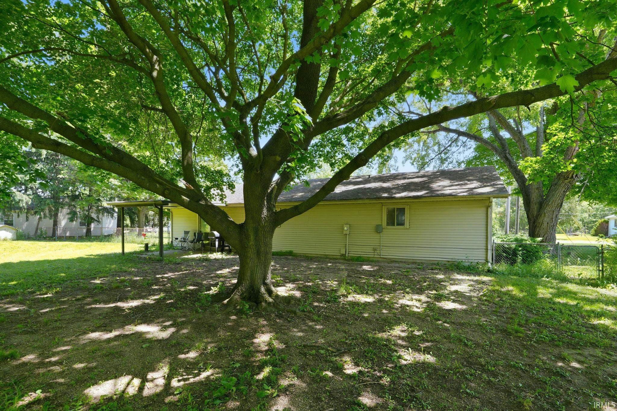 Expansive backyard featuring a mature tree providing shade, a detached structure with light-colored siding, a covered patio area, a chain-link fence, and a grassy lawn