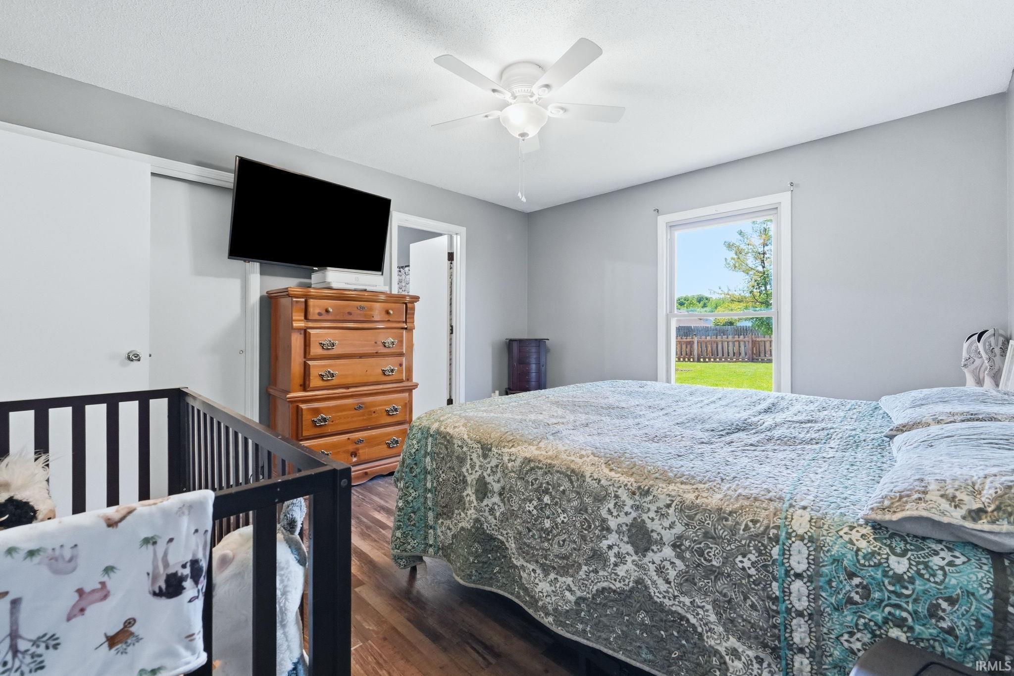 Bedroom featuring wood-finish flooring, light gray walls, a ceiling fan with light, and a window overlooking a fenced yard