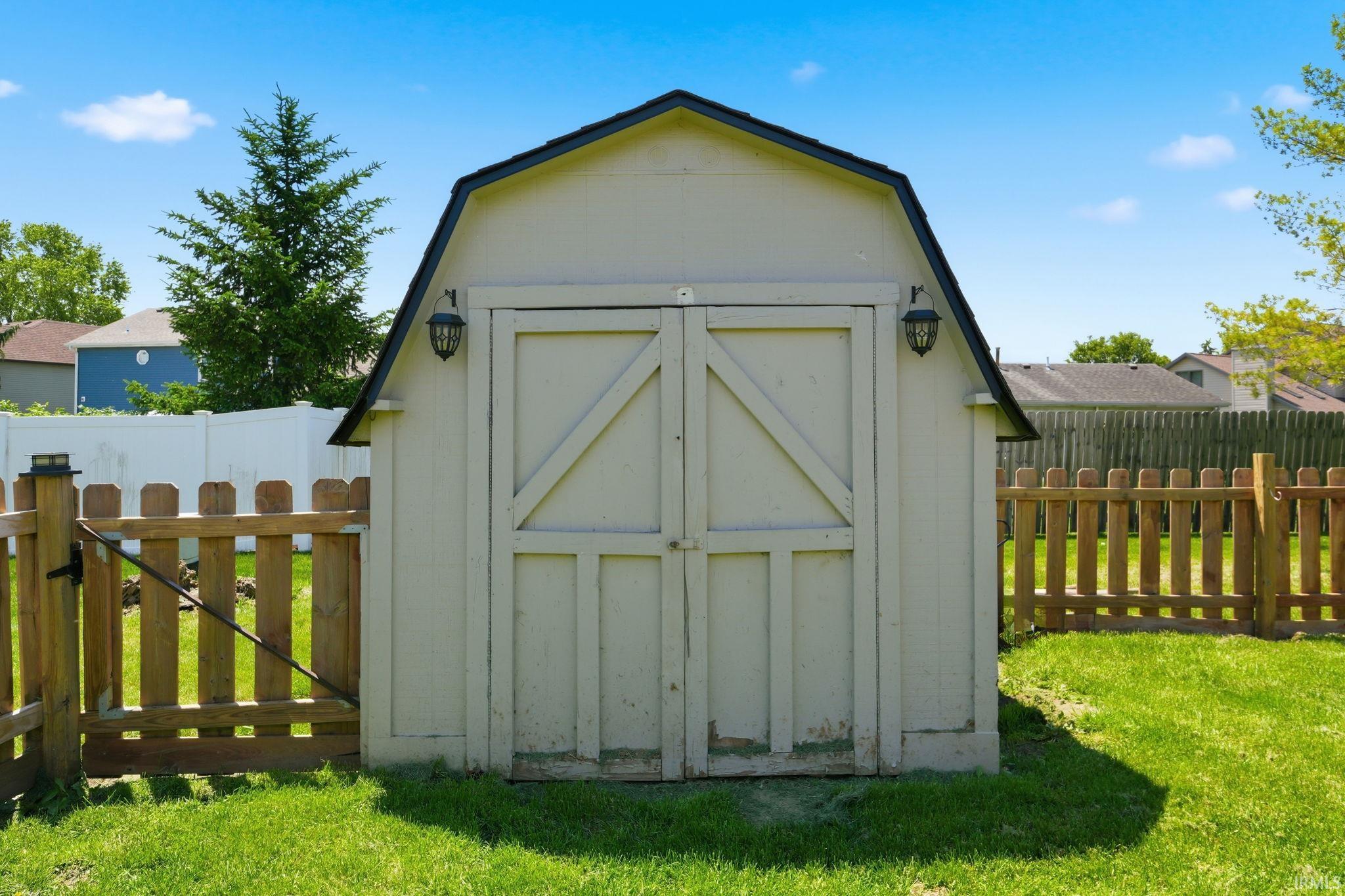 Gabled storage shed with double barn doors, exterior light fixtures, and perimeter fencing