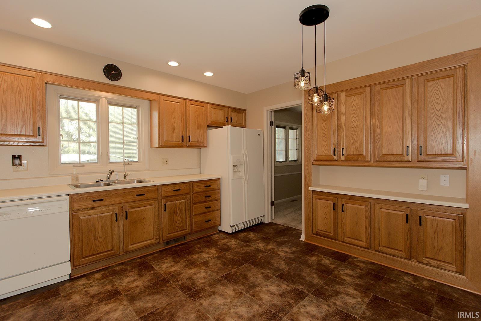 Kitchen featuring abundant wood cabinetry, white countertops, a double basin sink beneath a large window, a white refrigerator, and a three-light pendant fixture