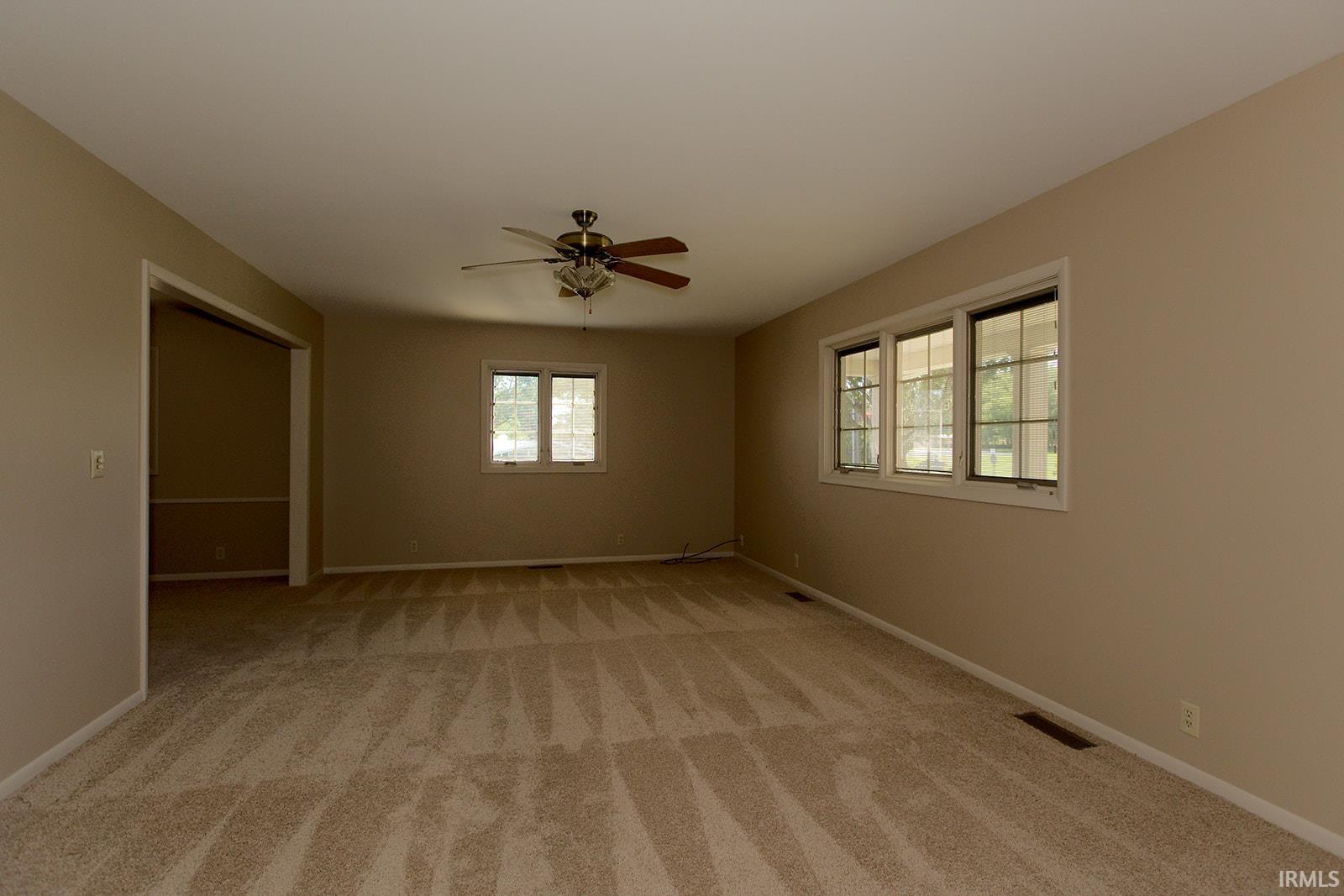 Spacious room featuring neutral carpeting, light-toned walls, multiple windows providing natural illumination, and a ceiling fan with integrated lighting