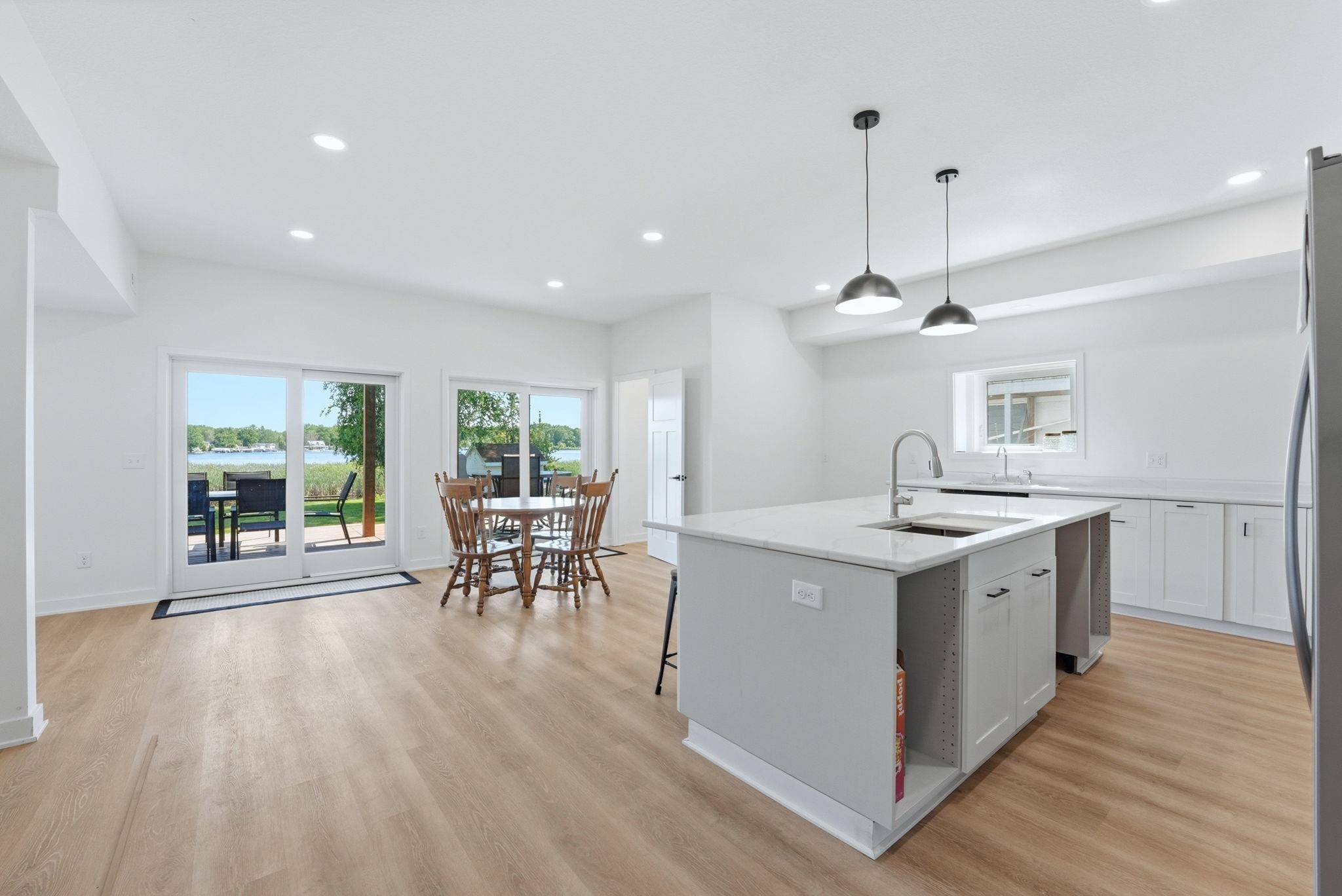 Open-concept kitchen and dining area featuring light wood-finish flooring, white cabinetry with dark hardware, a center island with sink and pendant lighting, and sliding glass doors opening to a body of water