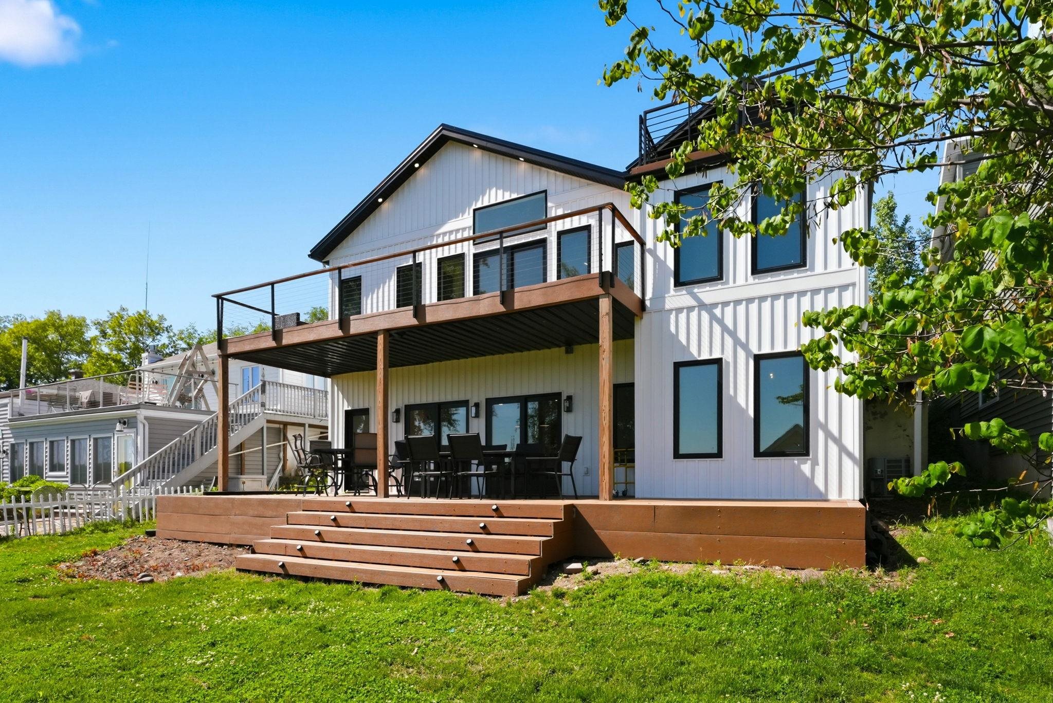 Modern exterior featuring white vertical siding, black trim windows, a ground-level deck with integrated steps, and an upper-level balcony with glass railing