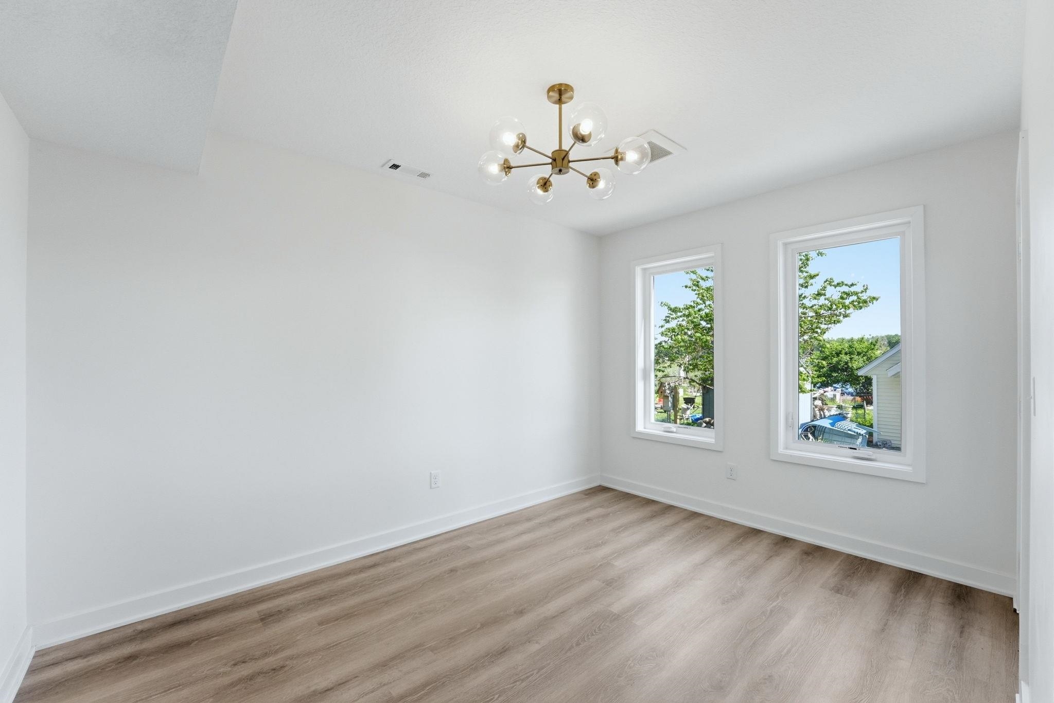 Bright interior space featuring a modern gold-finish chandelier, two large windows with white trim, wood-finish flooring, and white baseboards