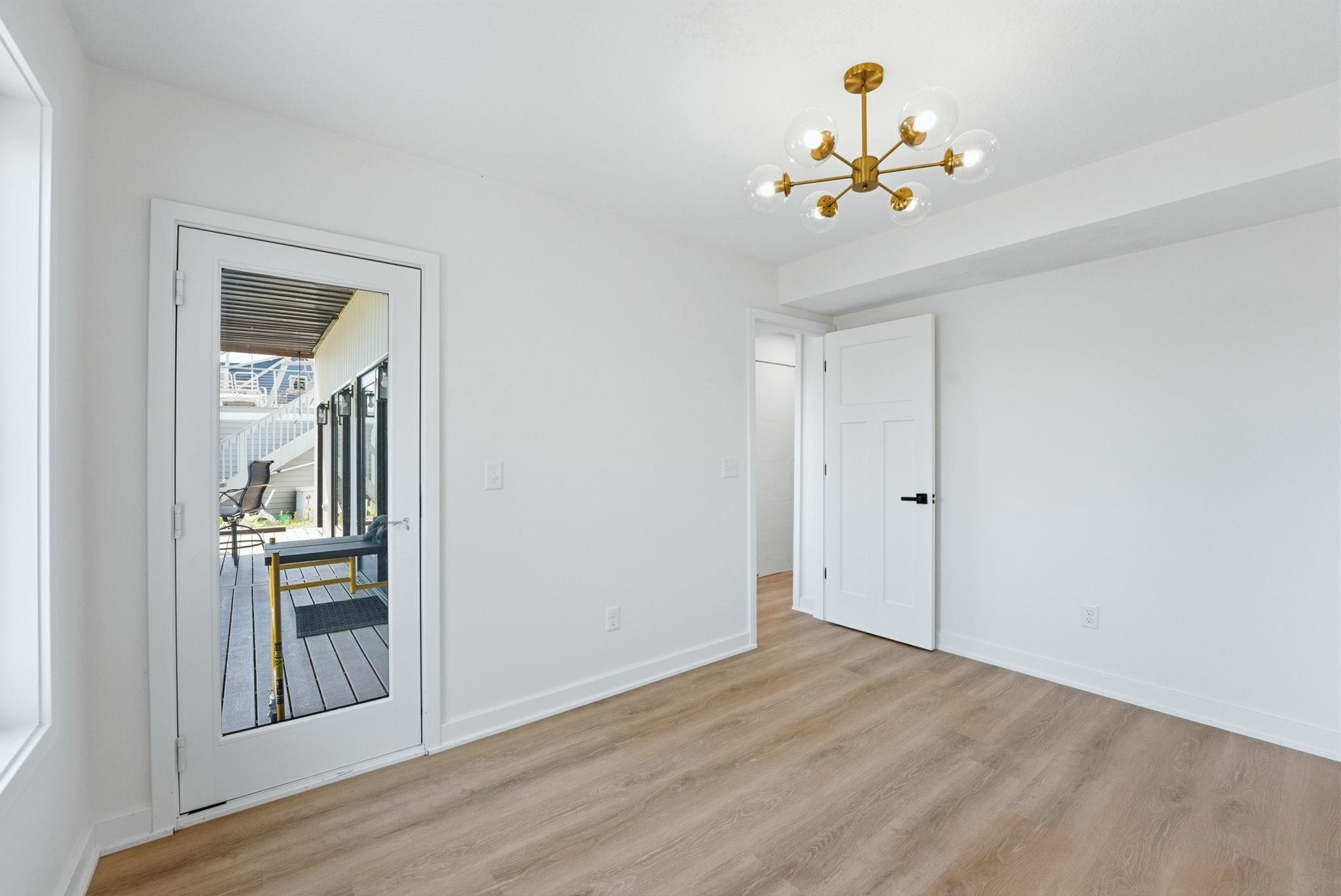 Bright interior space featuring wood-finish flooring, a modern brass and glass chandelier, a white paneled door with matte black hardware, and a glass-paned door opening to an exterior deck