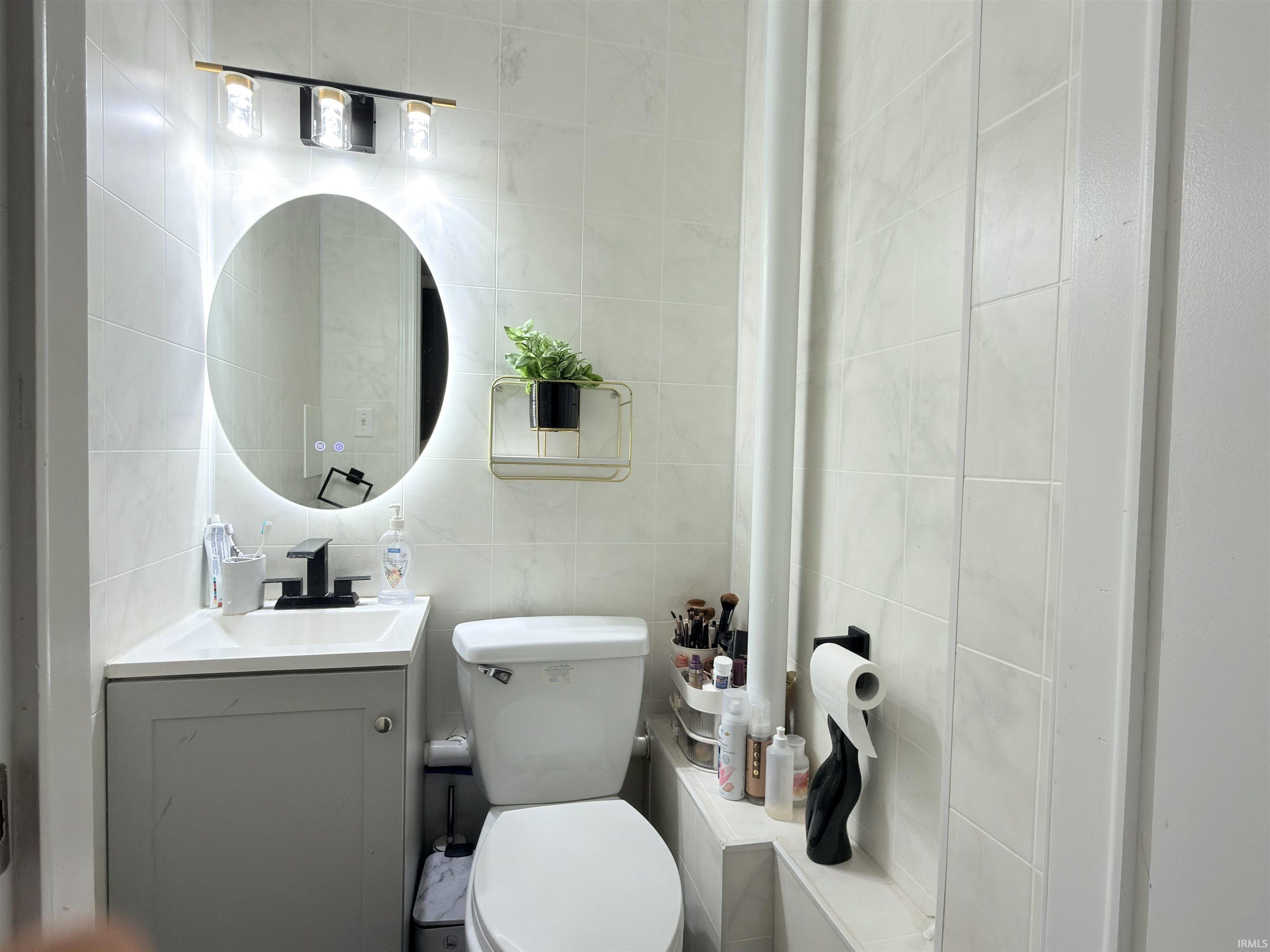 Bathroom featuring light-toned square wall tiles, a built-in vanity with a white countertop and basin, a matte black faucet, and an illuminated oval mirror