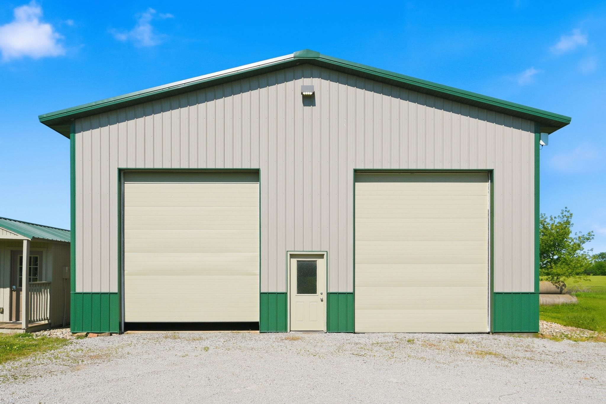 Two large garage doors on a metal-sided structure with green trim, a central entry door, and a gravel foreground
