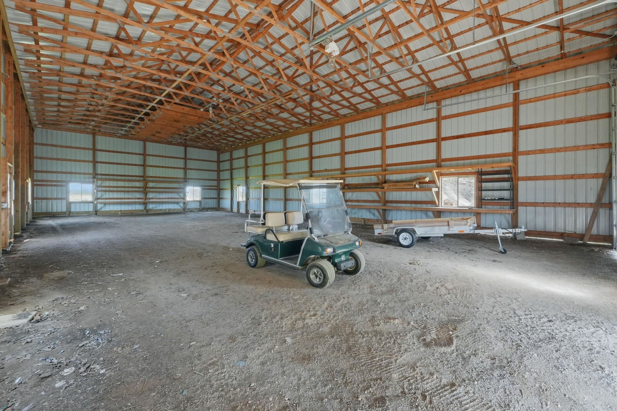 Spacious pole barn structure featuring exposed wood trusses, corrugated metal wall panels, multiple window openings, and an expansive gravel floor