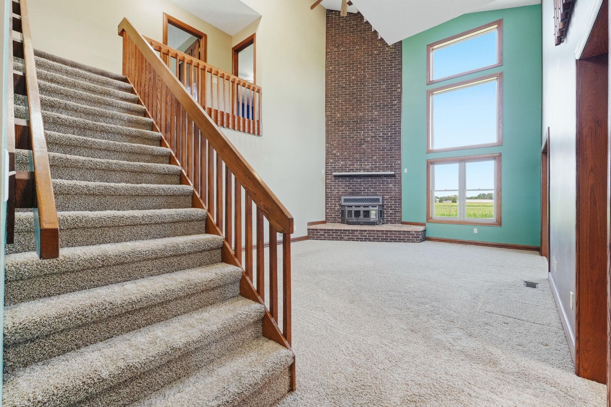Two-story living area featuring a prominent brick fireplace with a hearth, triple-stacked windows, and a carpeted staircase with a wood railing
