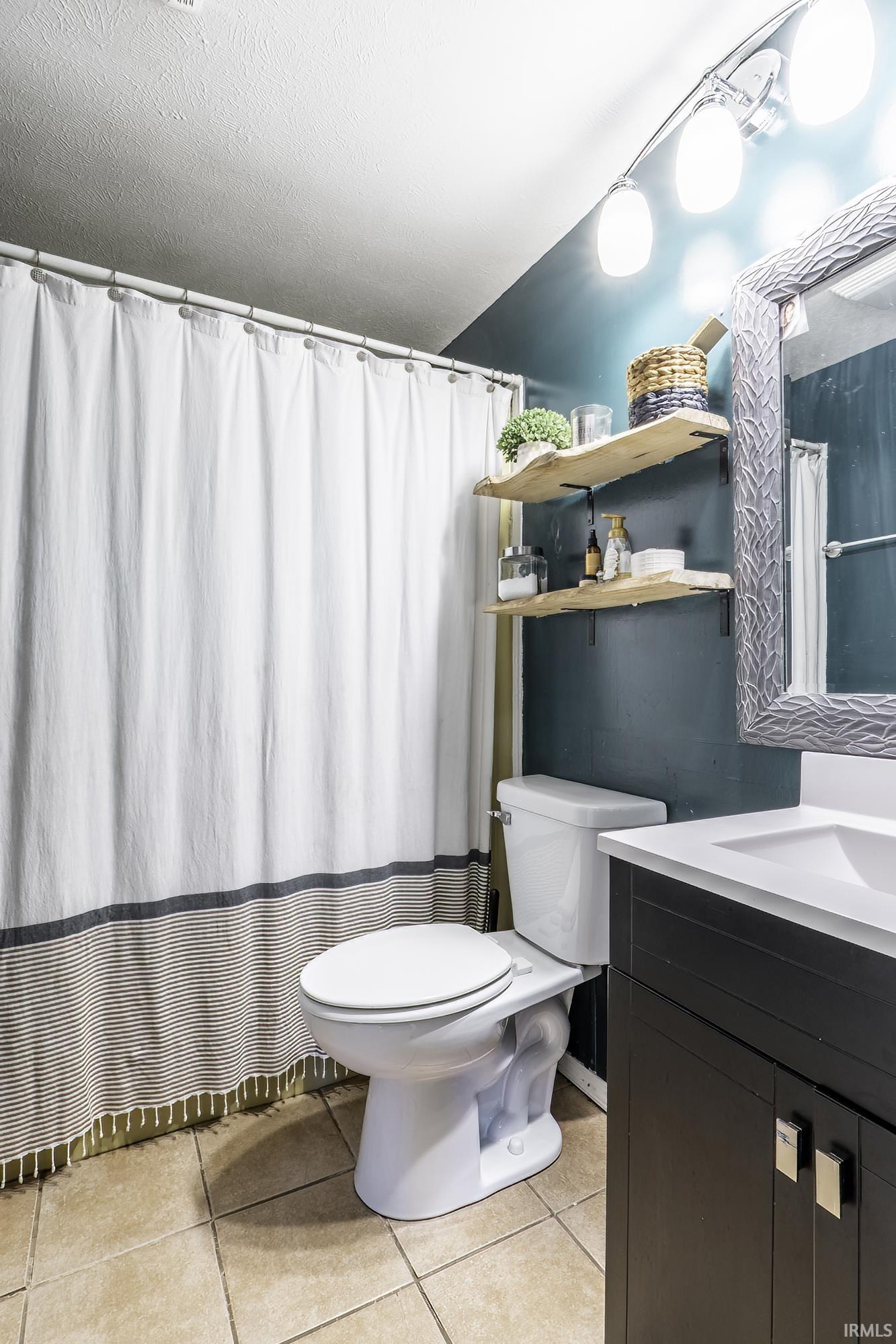 Bathroom featuring ceramic tile flooring, a dark vanity with a white countertop and integrated sink, a white toilet, and open wood shelving