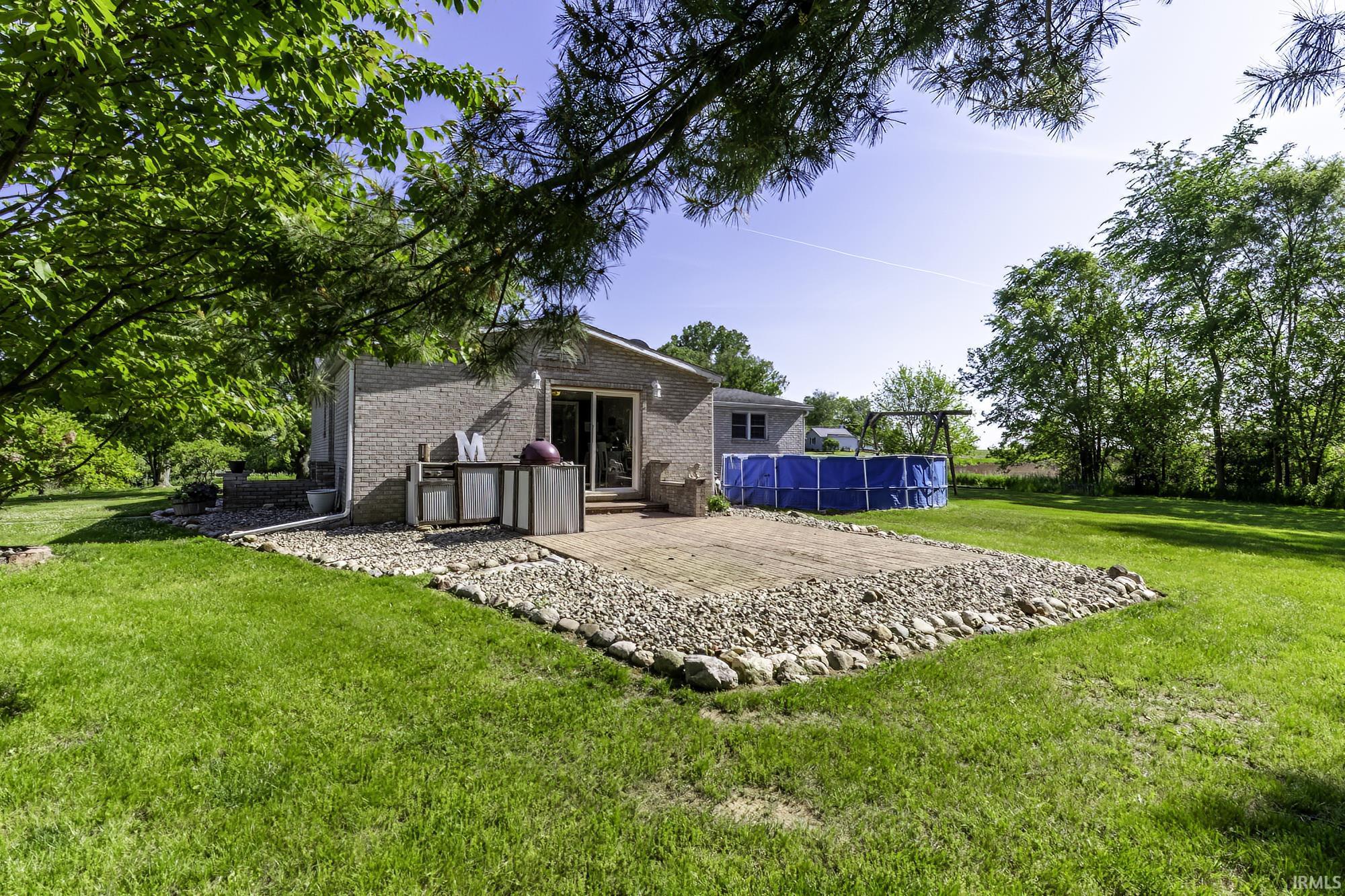 Expansive backyard featuring a stone patio with a river rock border, a blue above-ground pool, and mature trees lining the property perimeter
