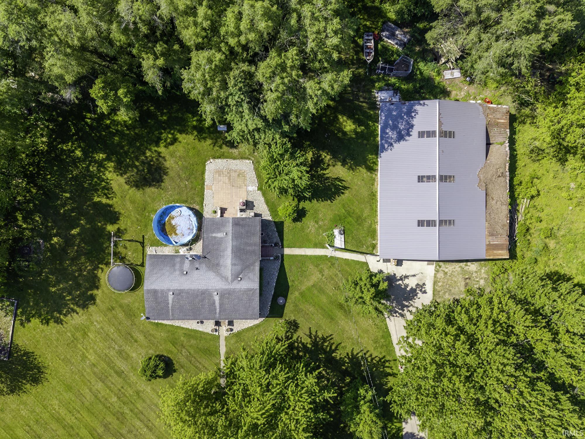 Expansive property featuring a large residential structure with a shingled roof, a substantial industrial-style building with a metal roof, mature surrounding trees, and a concrete pathway connecting the structures